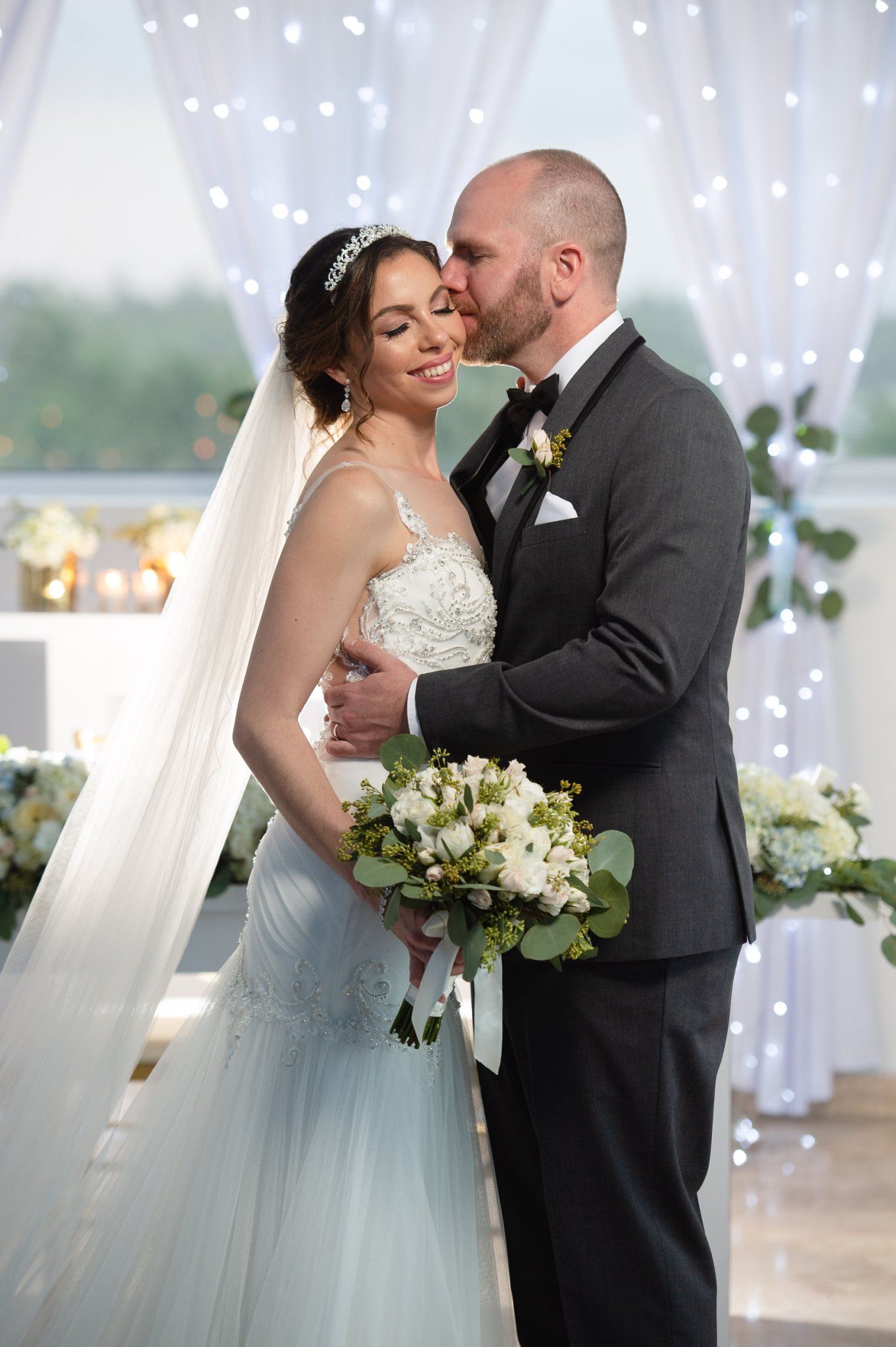 Bride and groom share a romantic kiss at wedding ceremony, bride holding white floral bouquet wearing lace gown and veil.