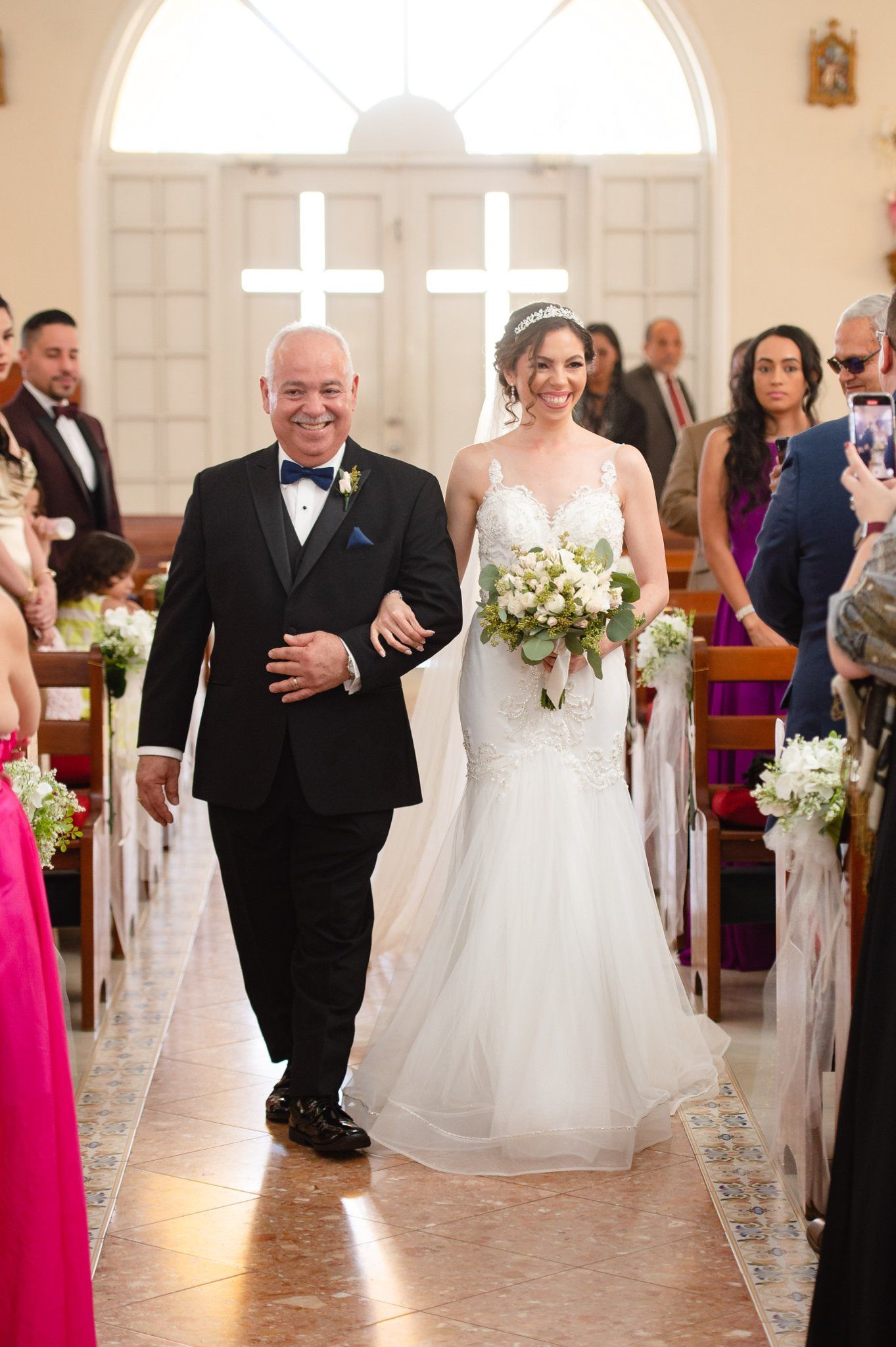 A bride in a white mermaid gown walks down the church aisle with her father in a black tuxedo holding a bouquet.