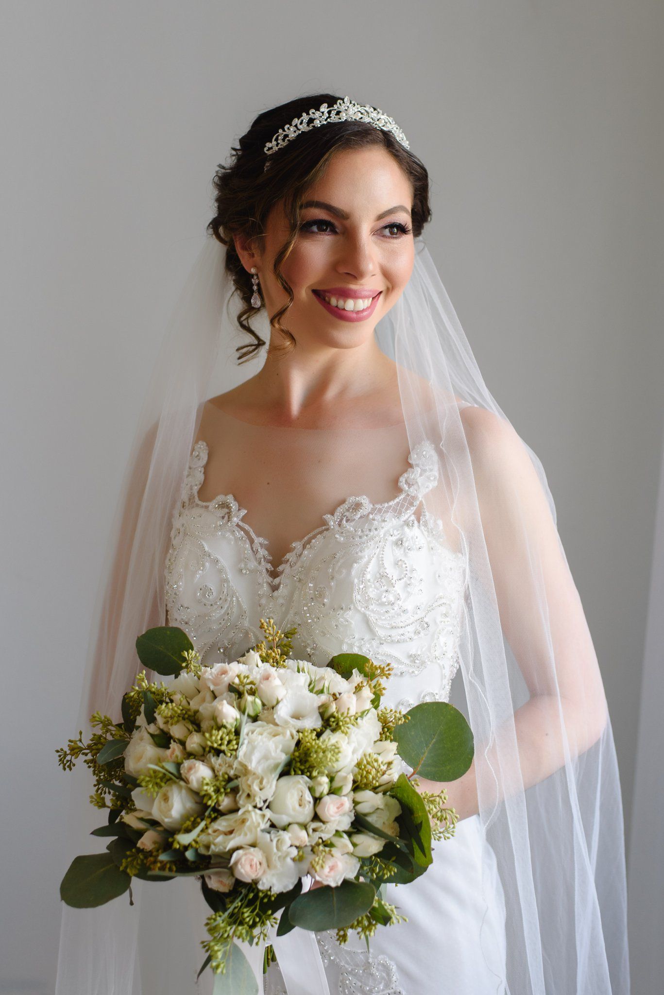 Beautiful bride smiling in lace wedding gown, crystal tiara, and veil while holding white and blush rose bouquet.