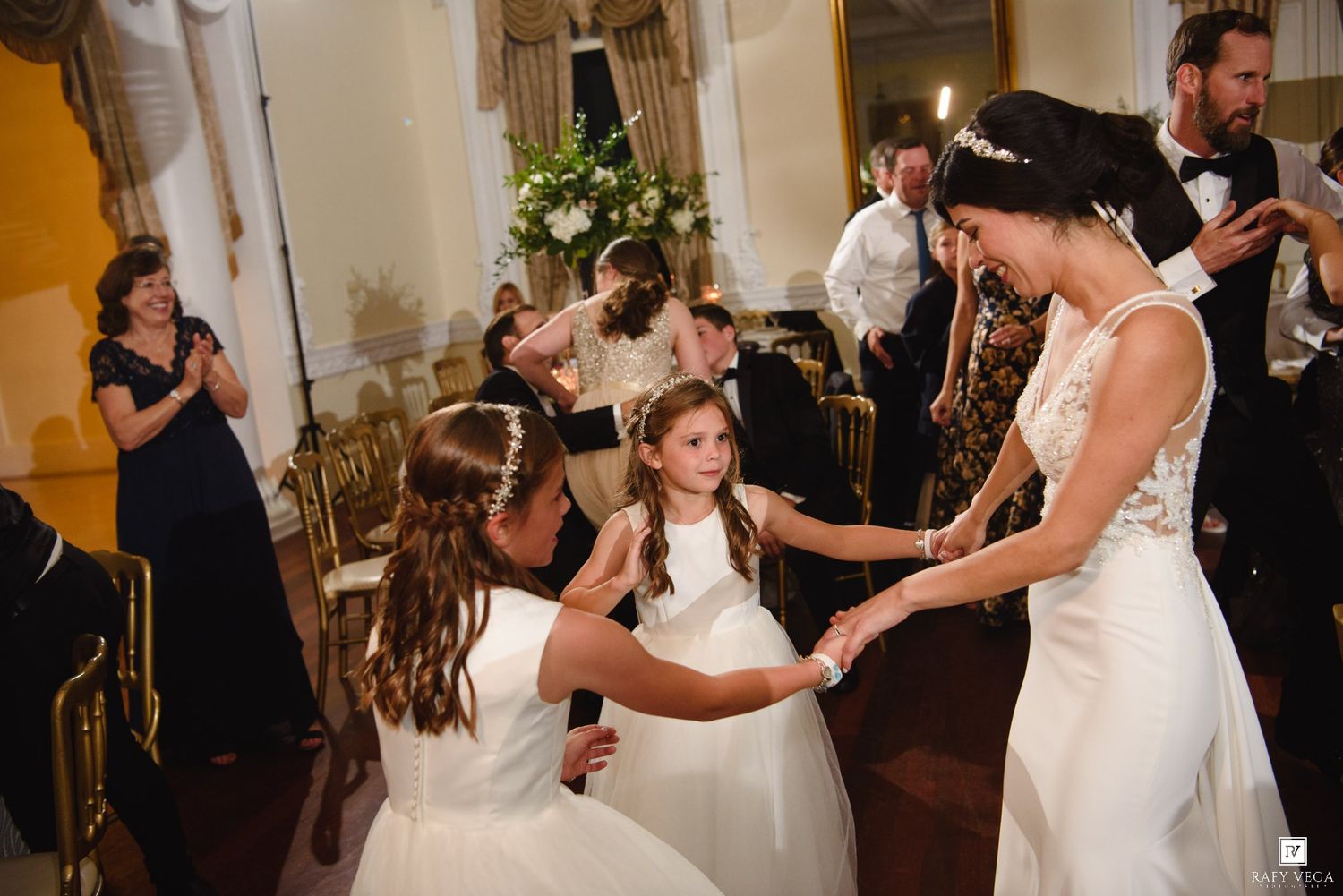 Bride in white gown dancing joyfully with flower girls in white dresses at elegant wedding reception.