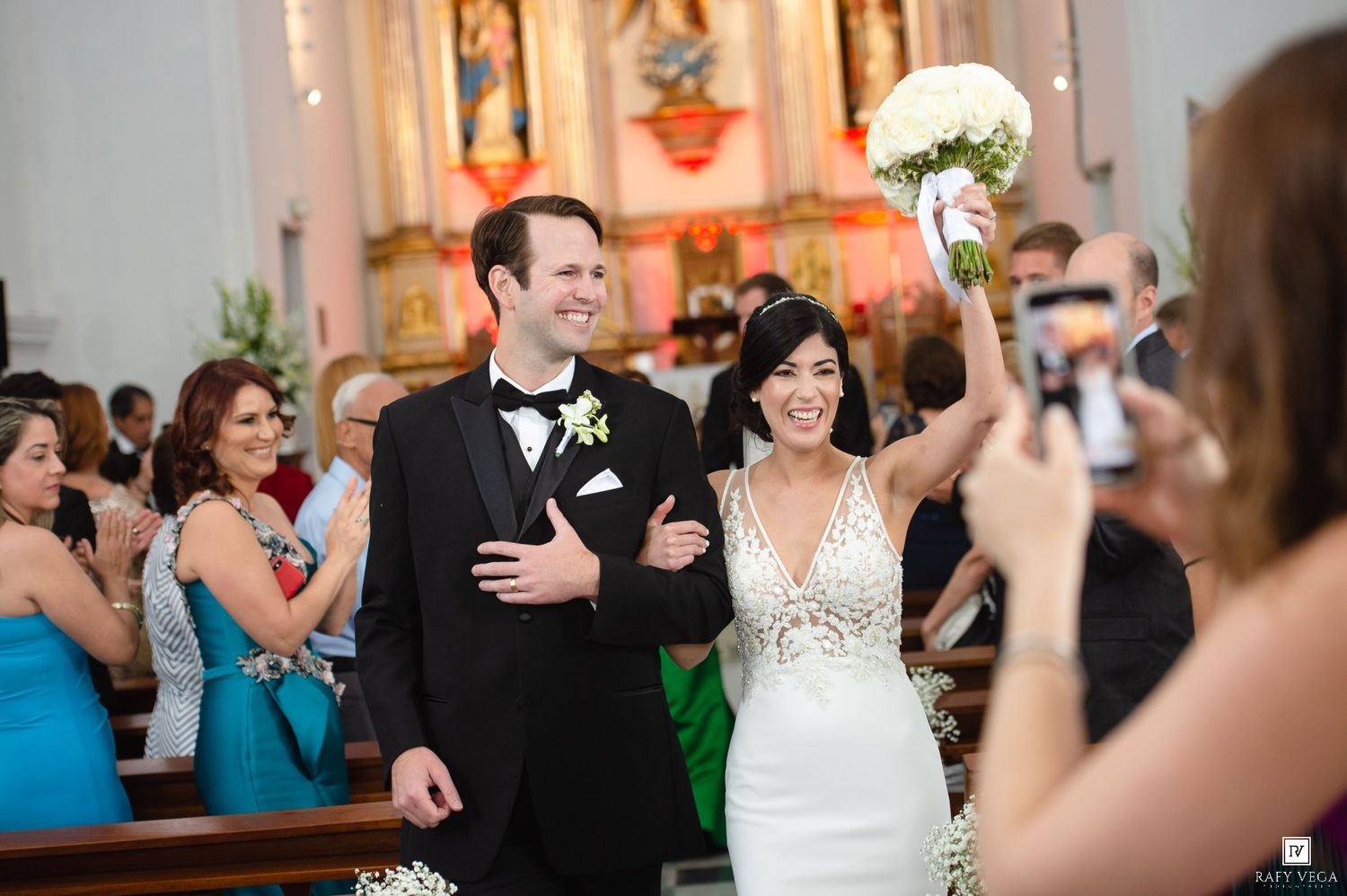 Joyful newlyweds walk down church aisle, bride raises bouquet triumphantly in white gown as guests celebrate wedding ceremony.