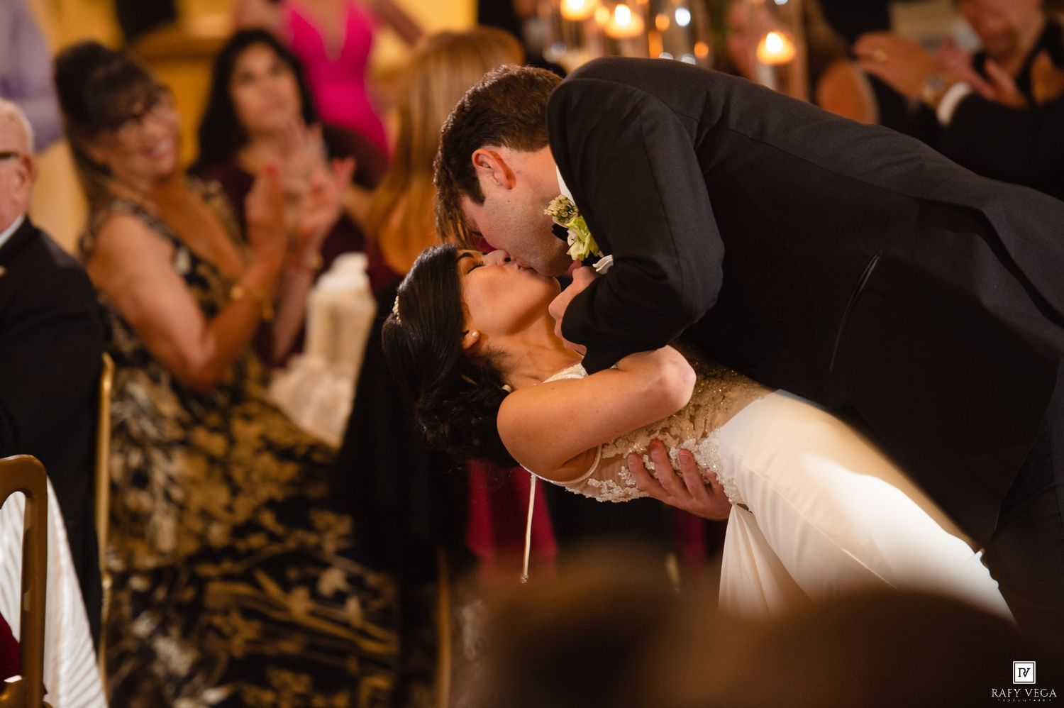 Bride and groom share a romantic dip kiss at their wedding reception while guests applaud in the background.