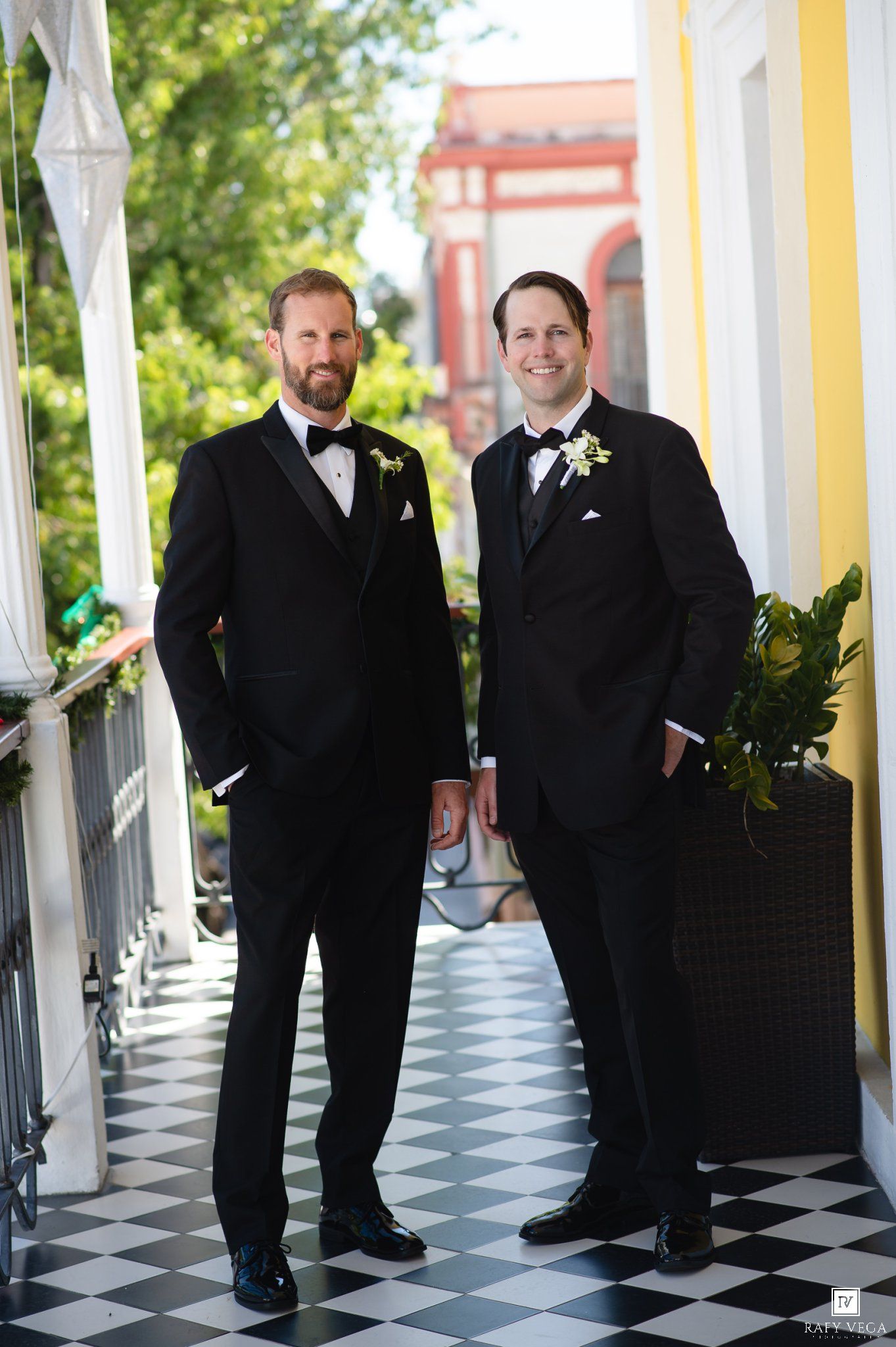 Two grooms in black tuxedos with boutonnieres pose on a checkered floor balcony at their wedding.