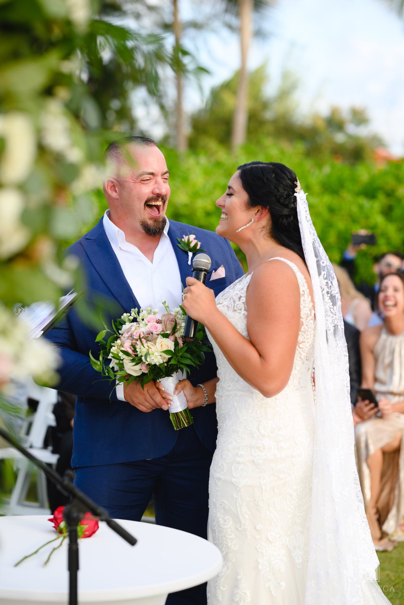 Bride and groom share a joyful moment during outdoor wedding ceremony, bride in lace gown holding pink bouquet.