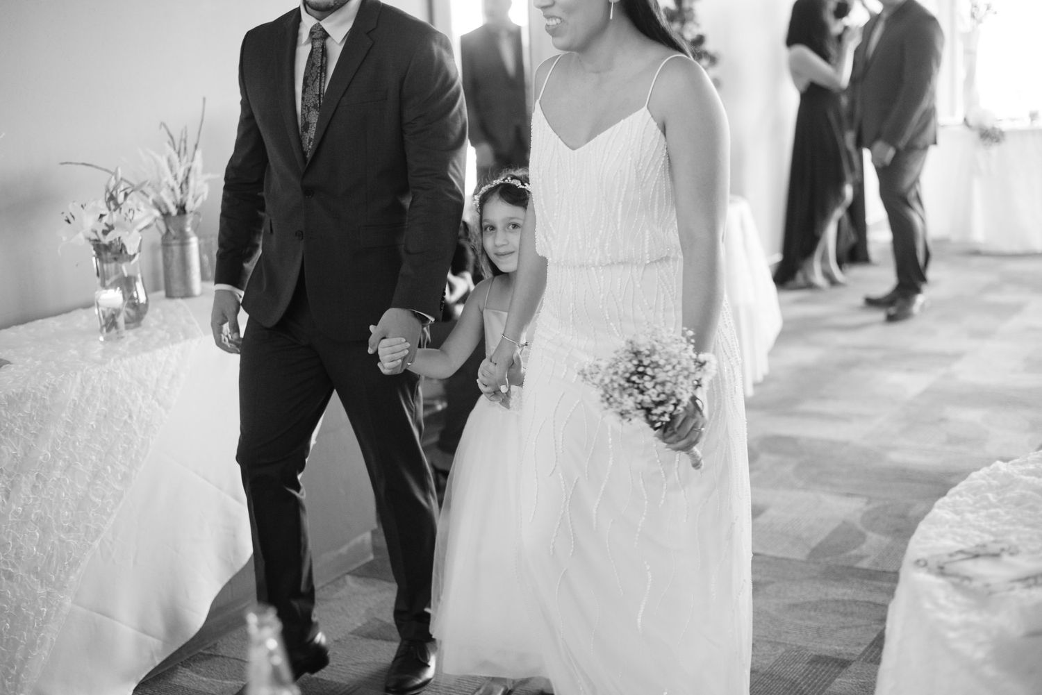 Black and white photo of bride and groom walking with flower girl at wedding reception.