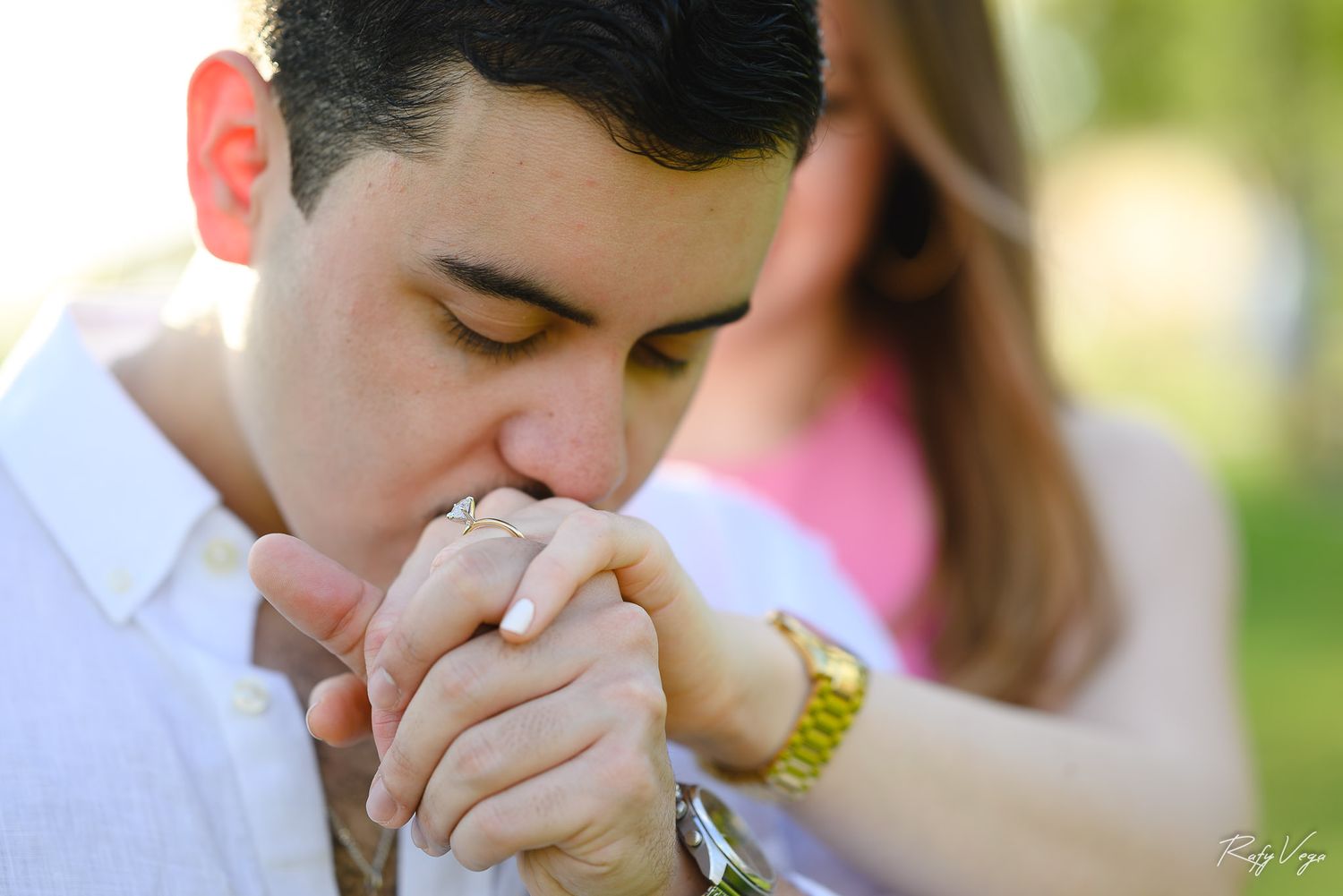 A man wearing a gold watch kisses a woman's hand tenderly during a romantic outdoor engagement photo session.