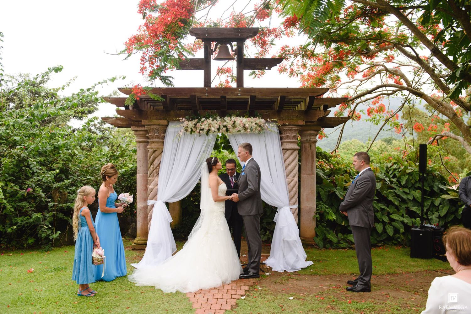 A bride and groom exchange vows under a wooden arch with white drapes and floral decor in a tropical garden wedding.