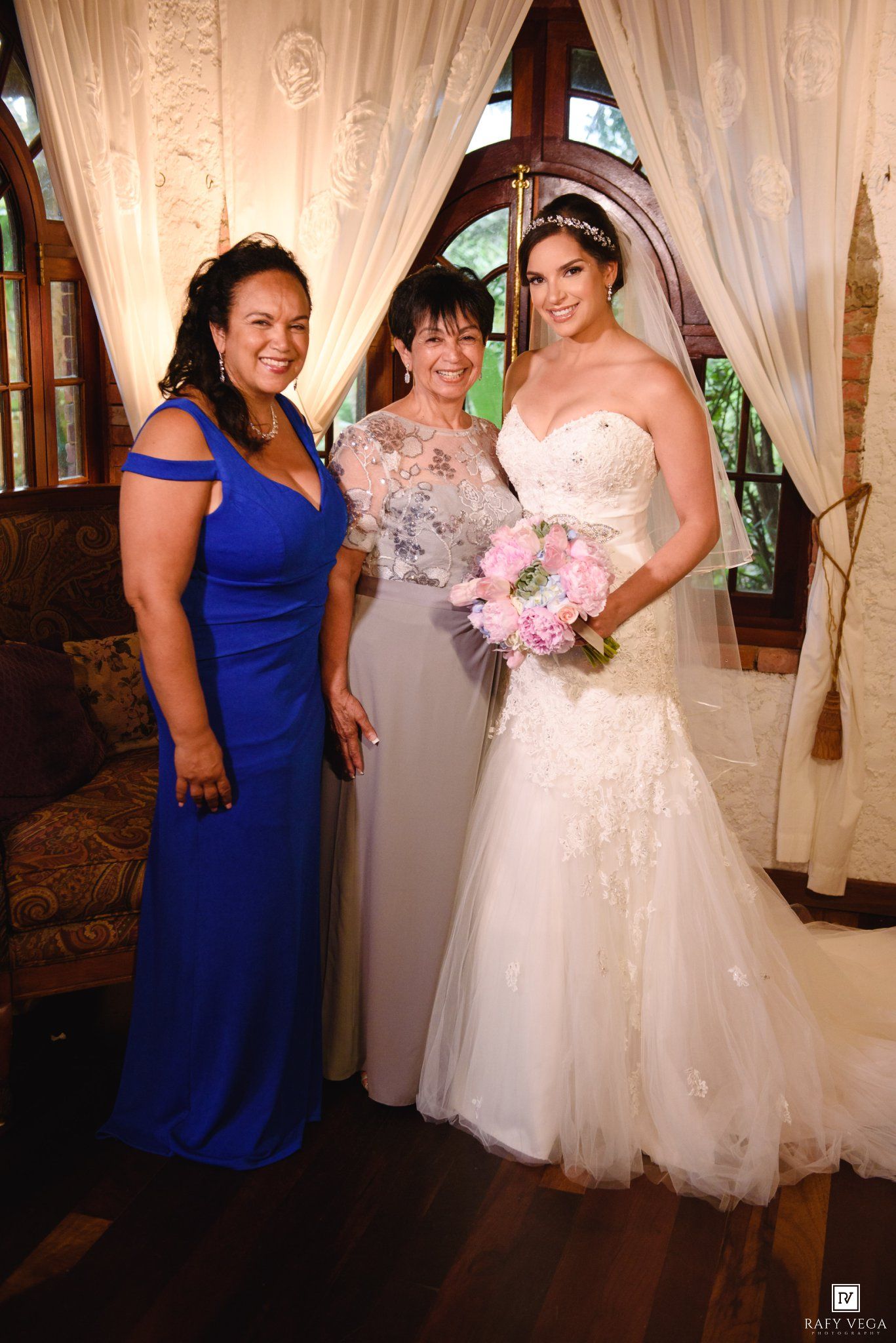 Bride in white gown holds pink bouquet, posing with two women in blue and gray dresses at a wedding venue.