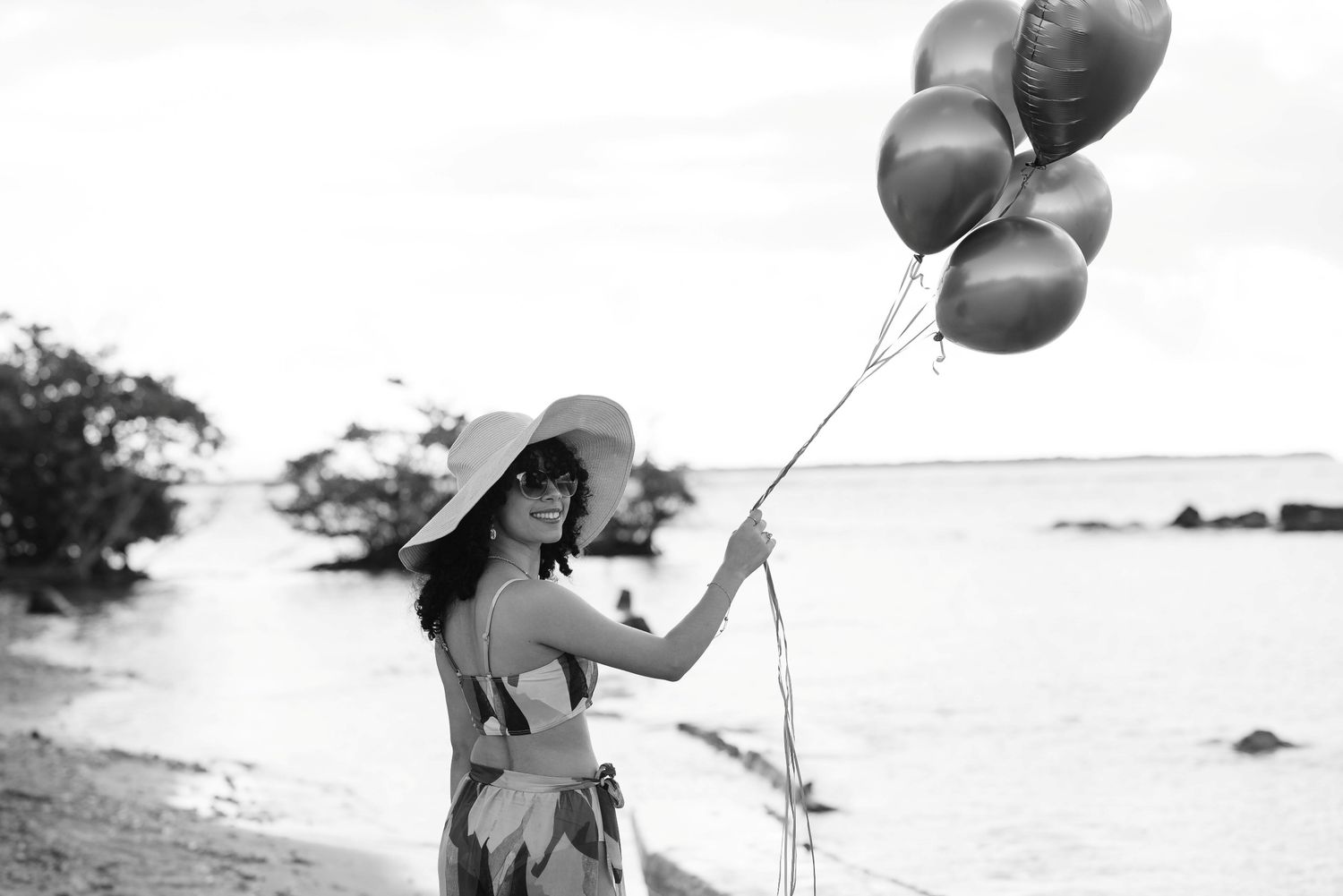 Woman in wide-brim hat holding balloons on a beach in black and white photo.