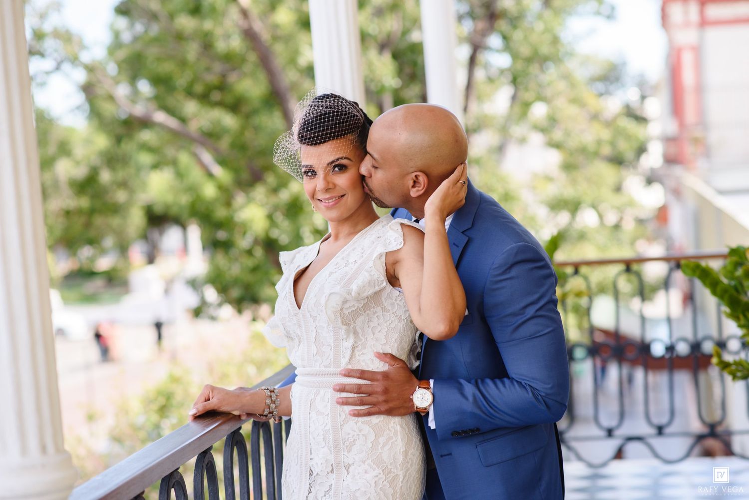 A groom in a blue suit kisses his bride in a white lace gown on a elegant balcony surrounded by columns.