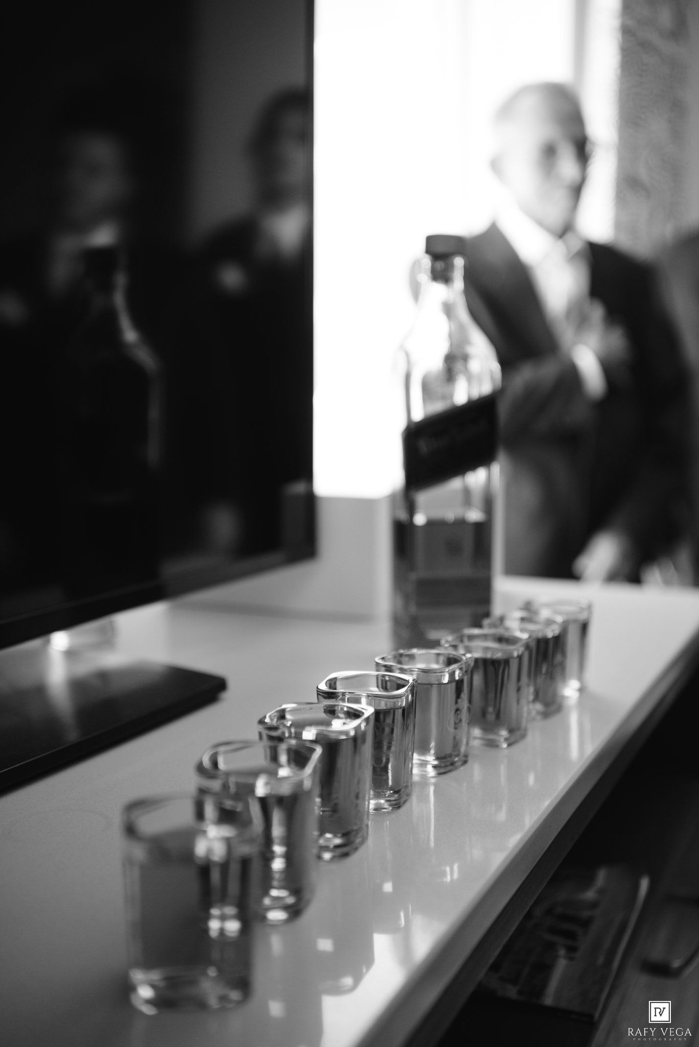 Row of shot glasses lined up on bar with bottle and groom in background at wedding reception.