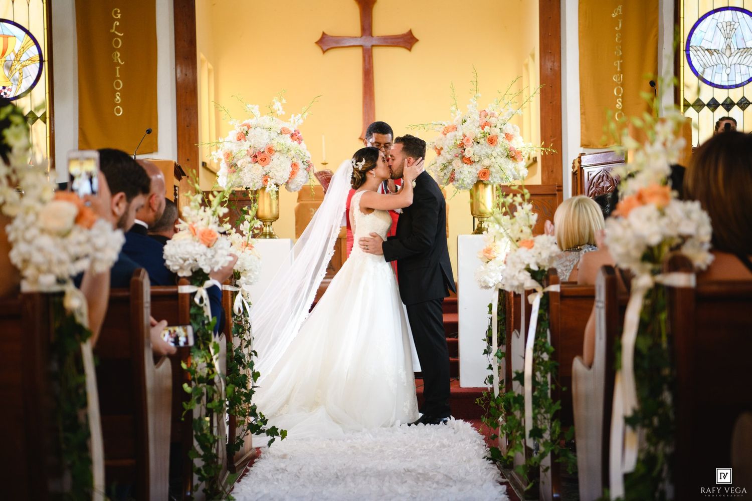 Bride and groom share first kiss at church altar adorned with white and peach floral arrangements during wedding ceremony.