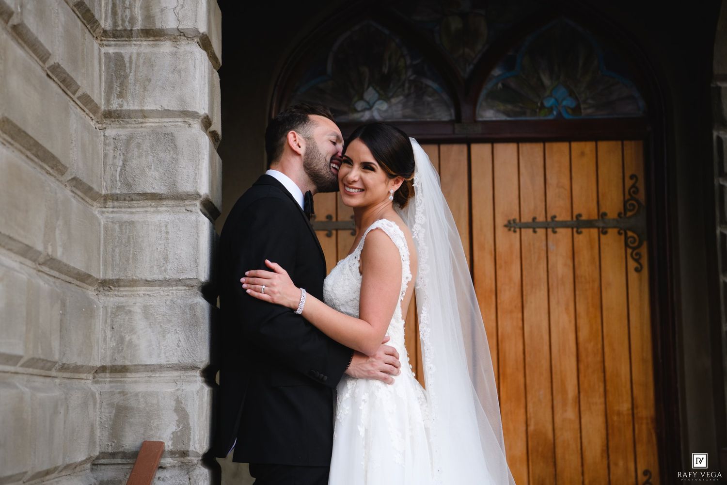 Groom kissing bride on cheek outside church doors, bride in white gown with veil, groom in black suit.