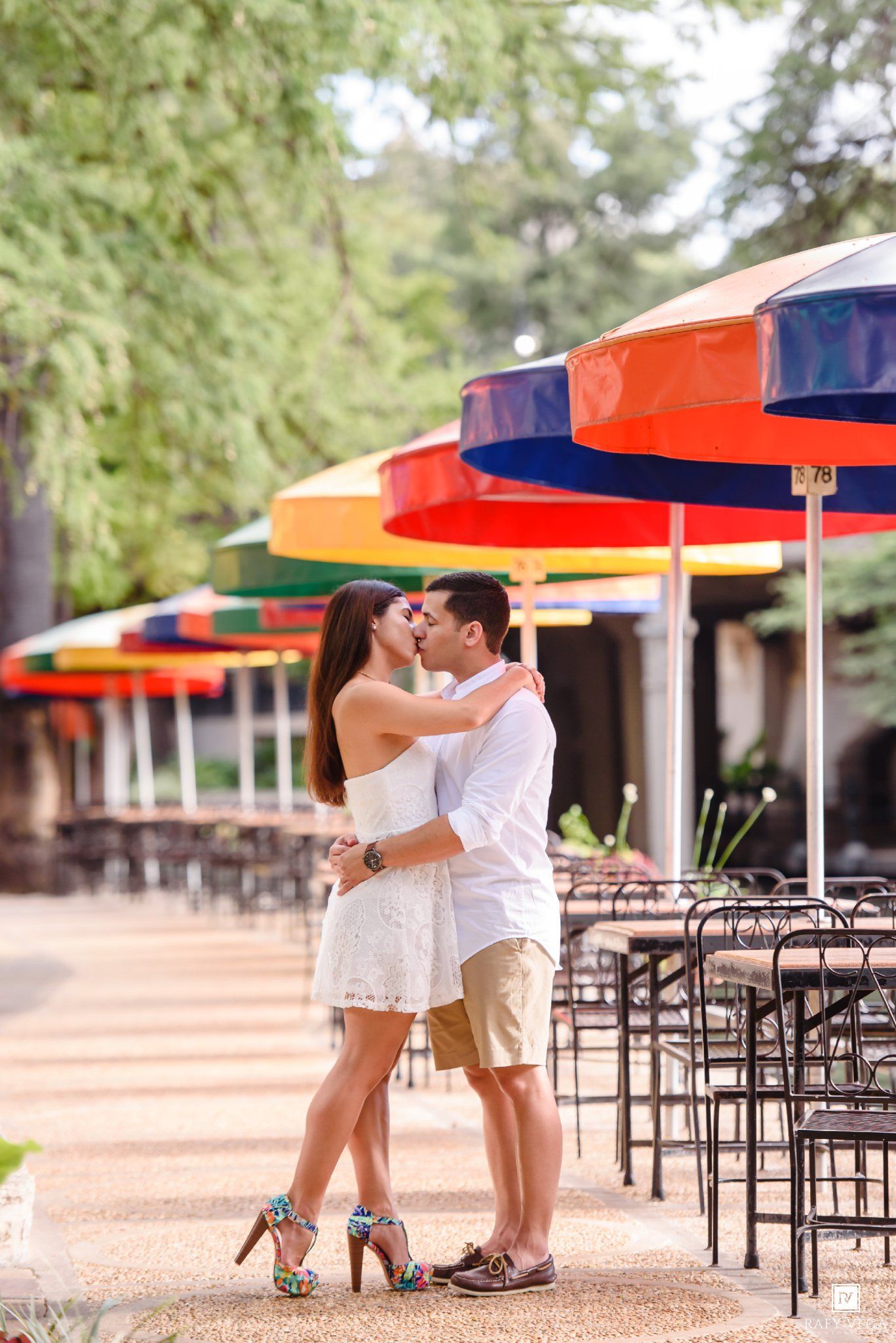 Couple sharing a romantic kiss near colorful outdoor umbrellas and cafe seating in a vibrant park setting.
