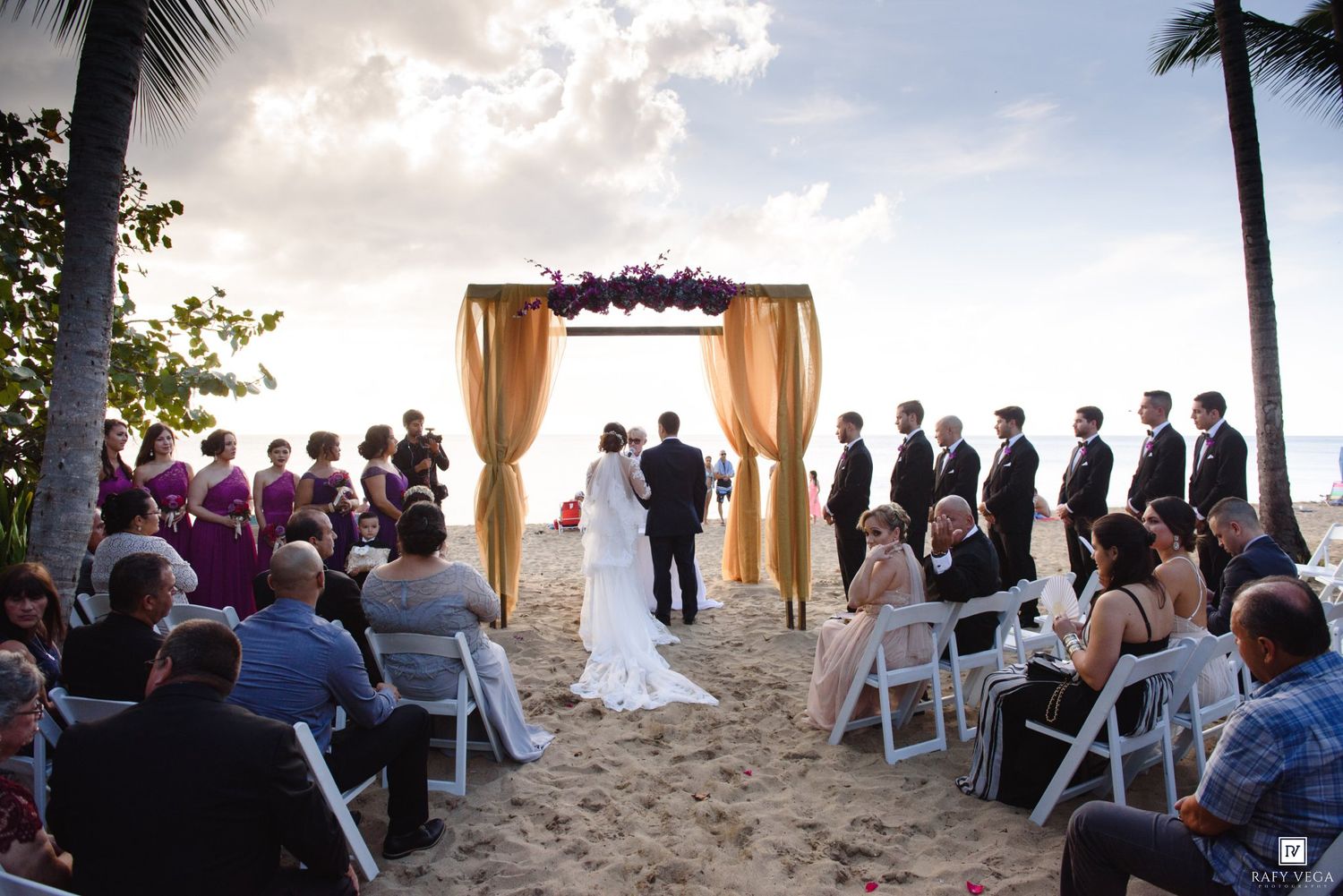 Romantic beach wedding ceremony with gold arch, bridal party, and guests seated on white chairs at sunset.