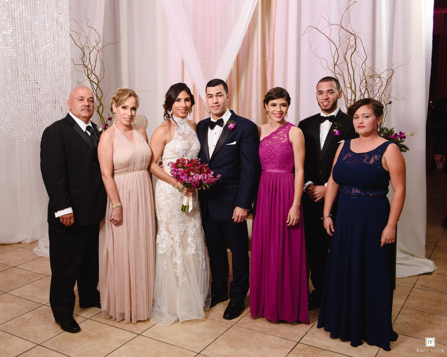 Wedding party poses formally in elegant venue, bride in white gown with bouquet, groom in tuxedo, flanked by family in formal attire.