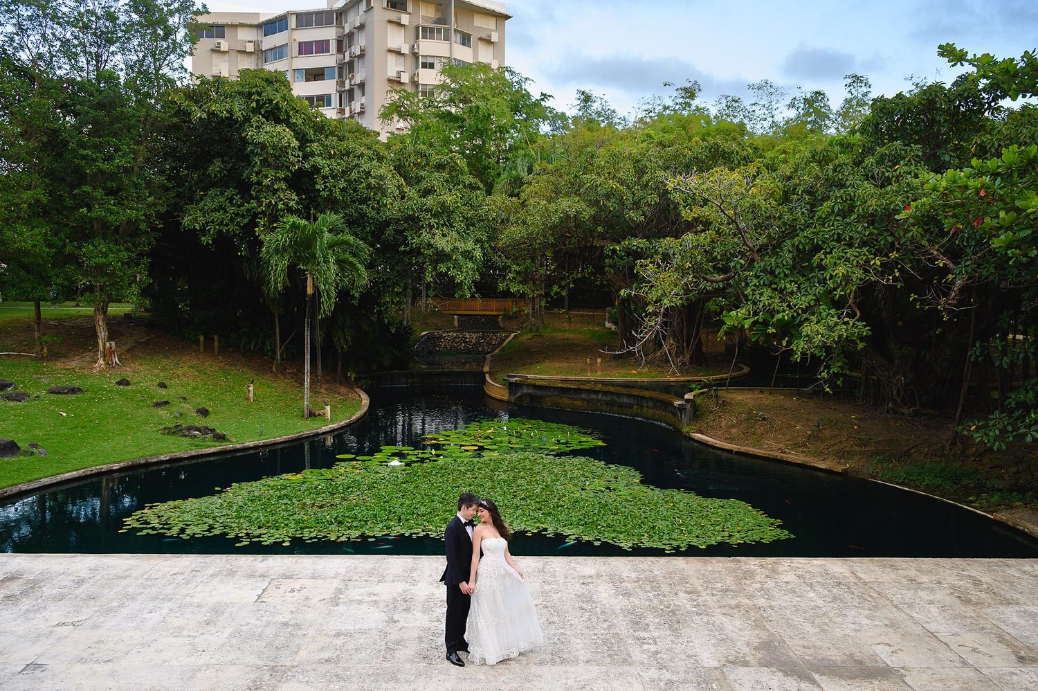 Bride and groom embracing near a lush green lily pond surrounded by tropical trees and a building backdrop.