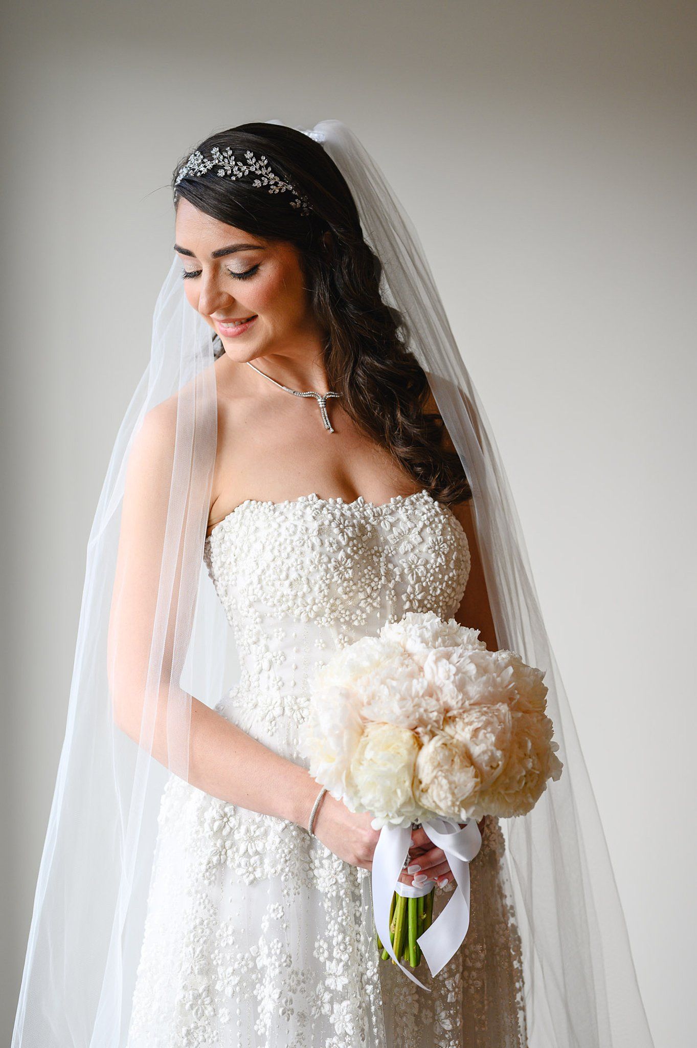 Bride in beaded strapless gown with veil and tiara holds white peony bouquet, smiling downward against neutral background.