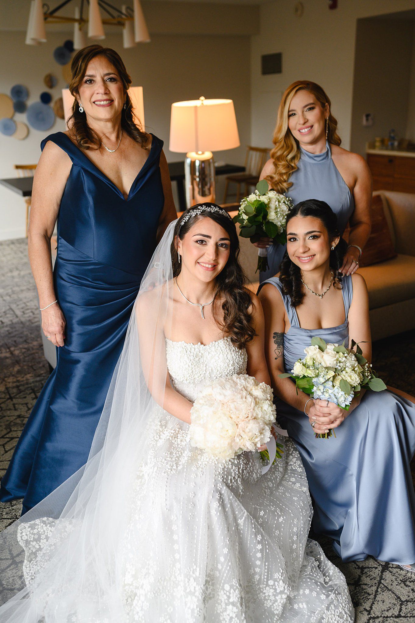 Bride in white gown with bridesmaids in blue dresses holding bouquets, posing together in elegant hotel room.