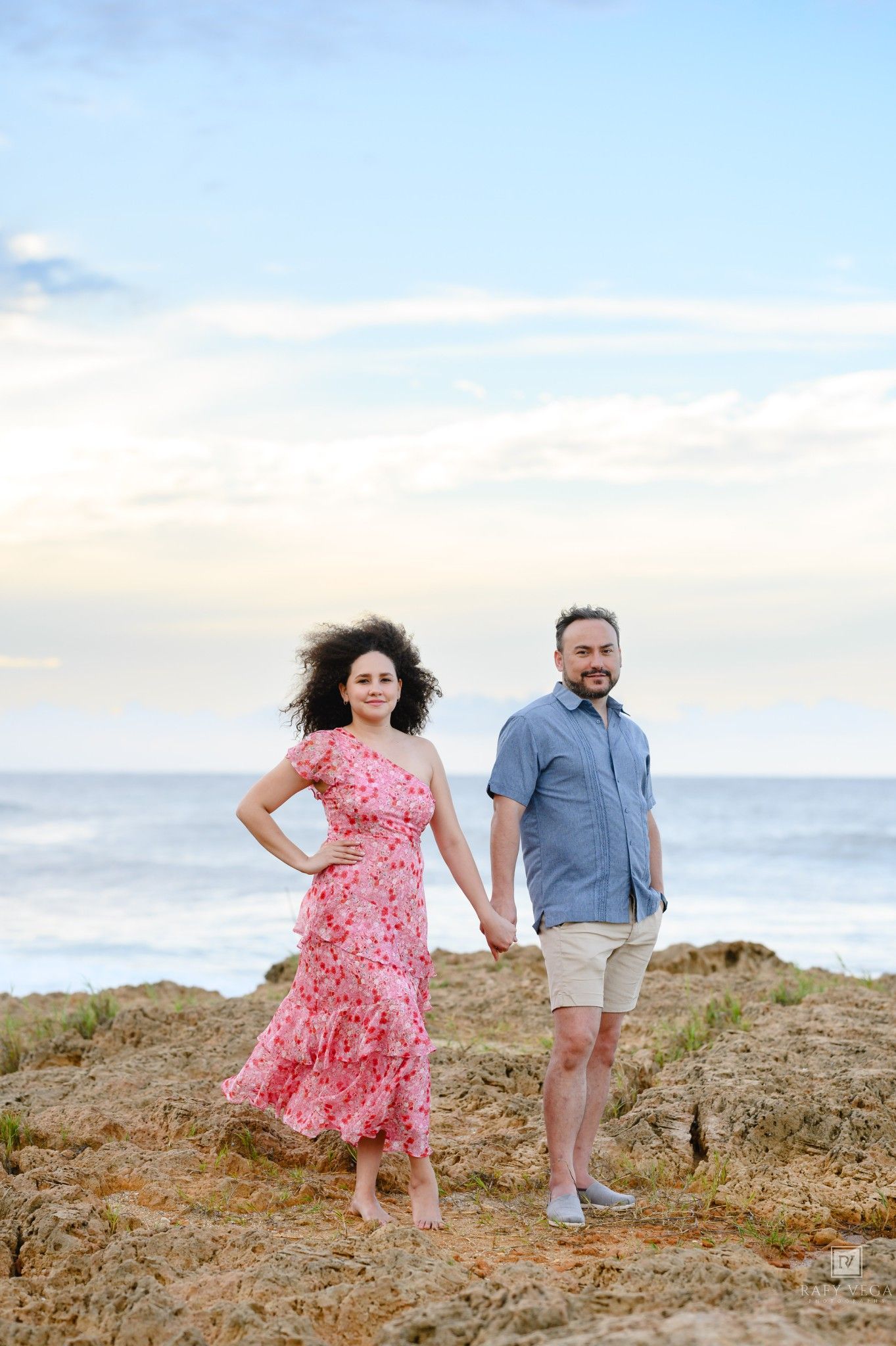 A couple holds hands on rocky coastal cliffs, woman in pink floral dress, man in blue shirt, ocean backdrop.