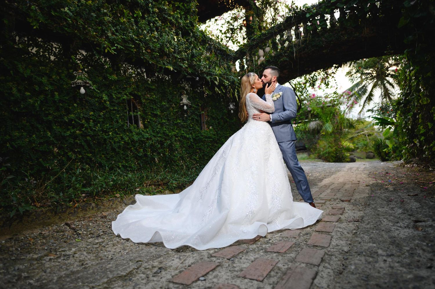 Bride in white ball gown and groom in gray suit embrace under ivy-covered stone bridge on brick path.