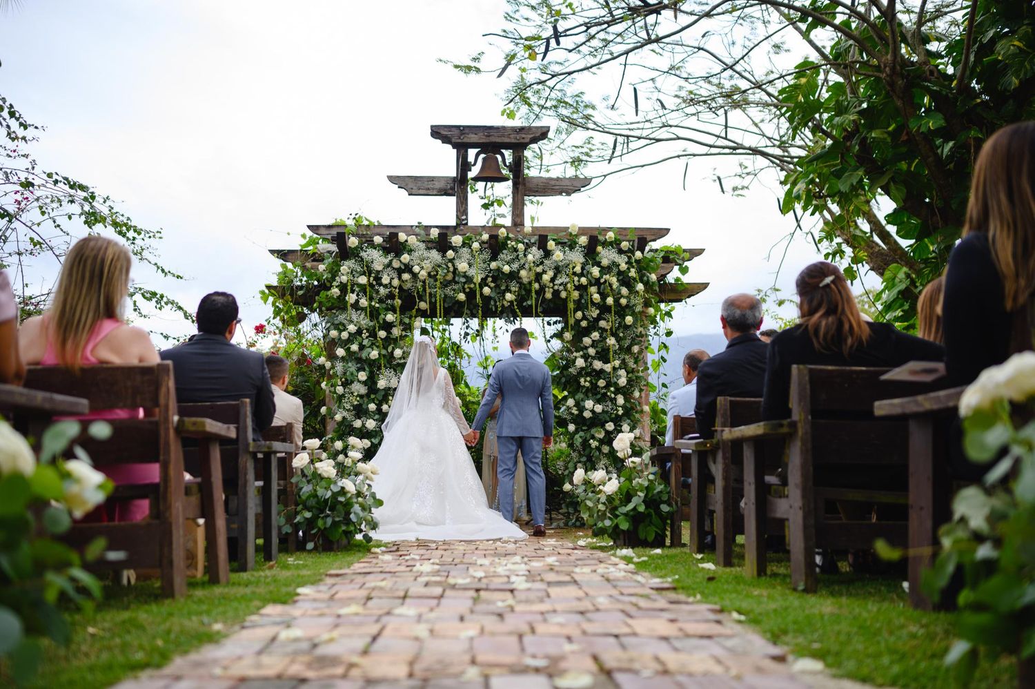Outdoor wedding ceremony with bride and groom at floral arch, guests seated on wooden pews in lush garden setting.