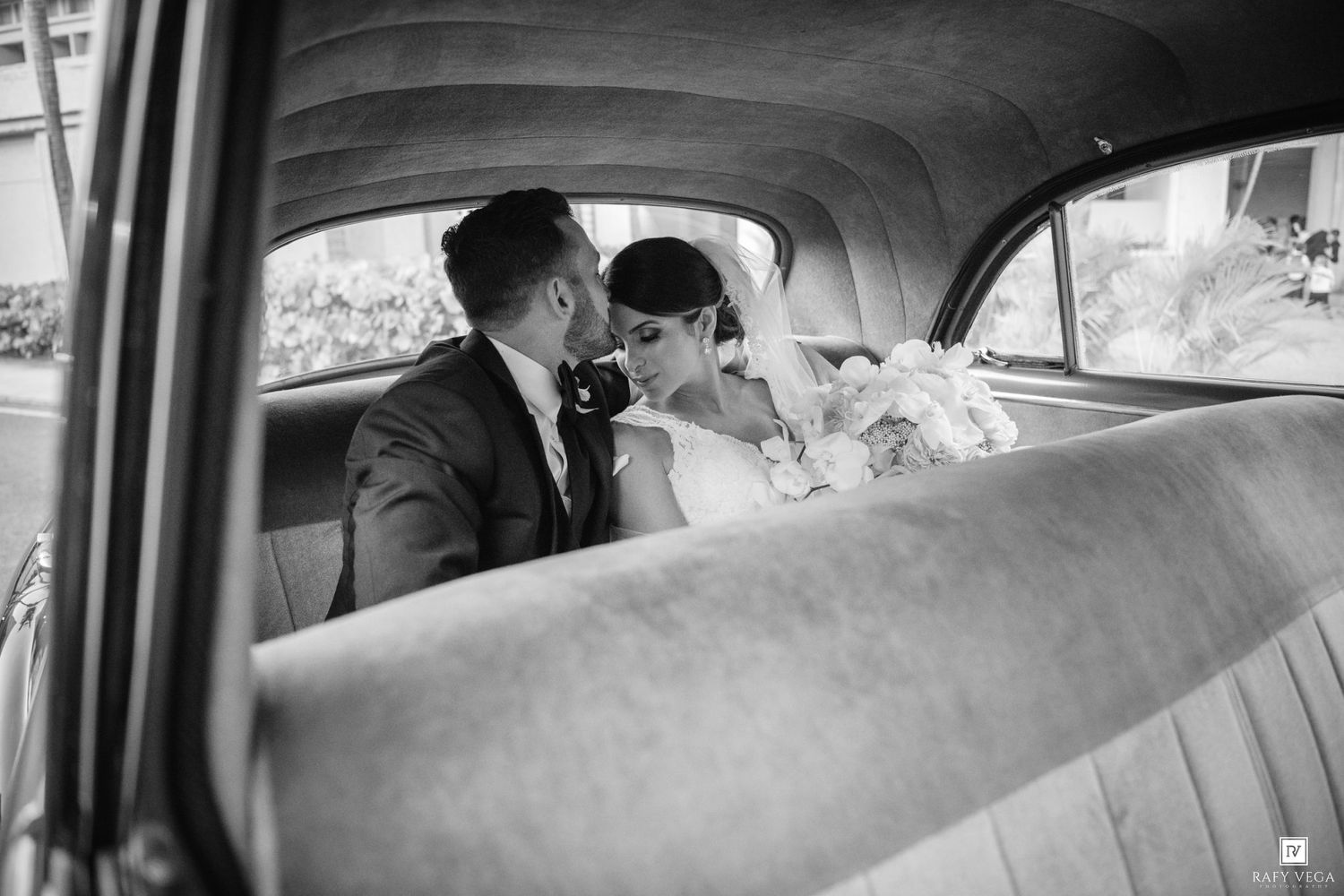 Black and white photo of a bride and groom sharing a romantic moment in the backseat of a vintage wedding car.