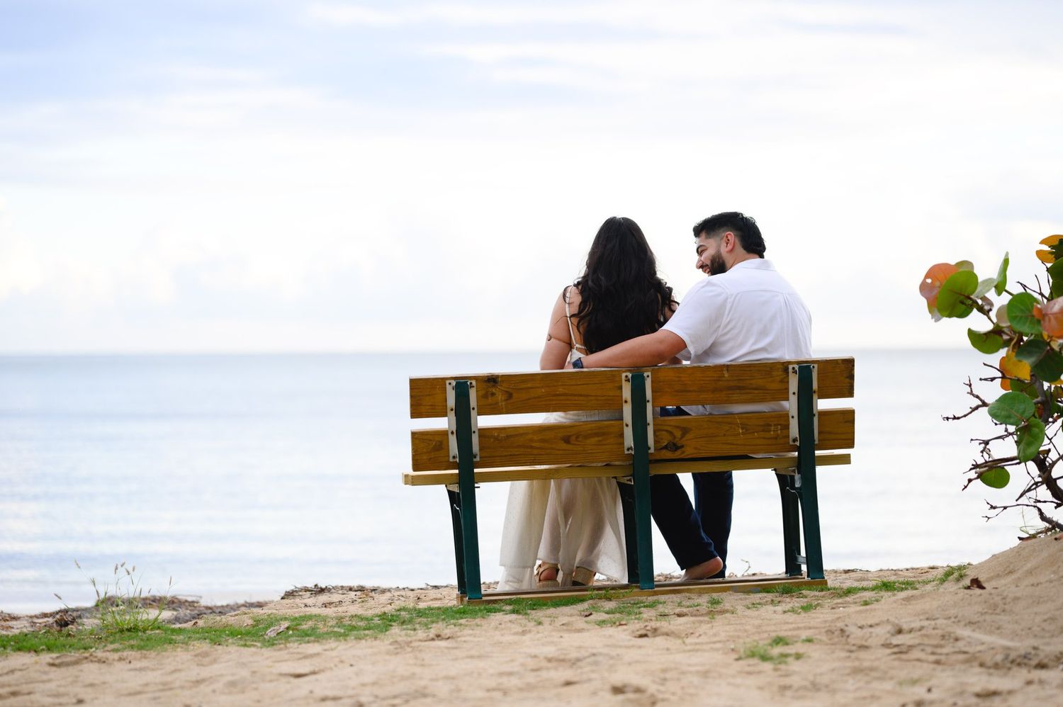 Couple sitting on a wooden bench overlooking a calm ocean on a sandy beach shore.