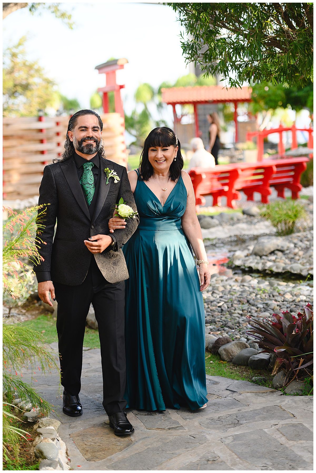 Couple in elegant teal gown and black suit walking through a garden venue on their wedding day.