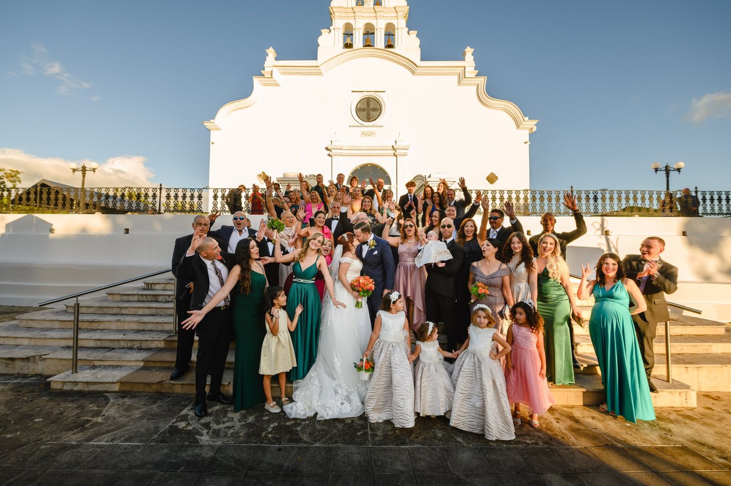Large wedding party celebrating in front of a white church, bridesmaids in green dresses, guests cheering.
