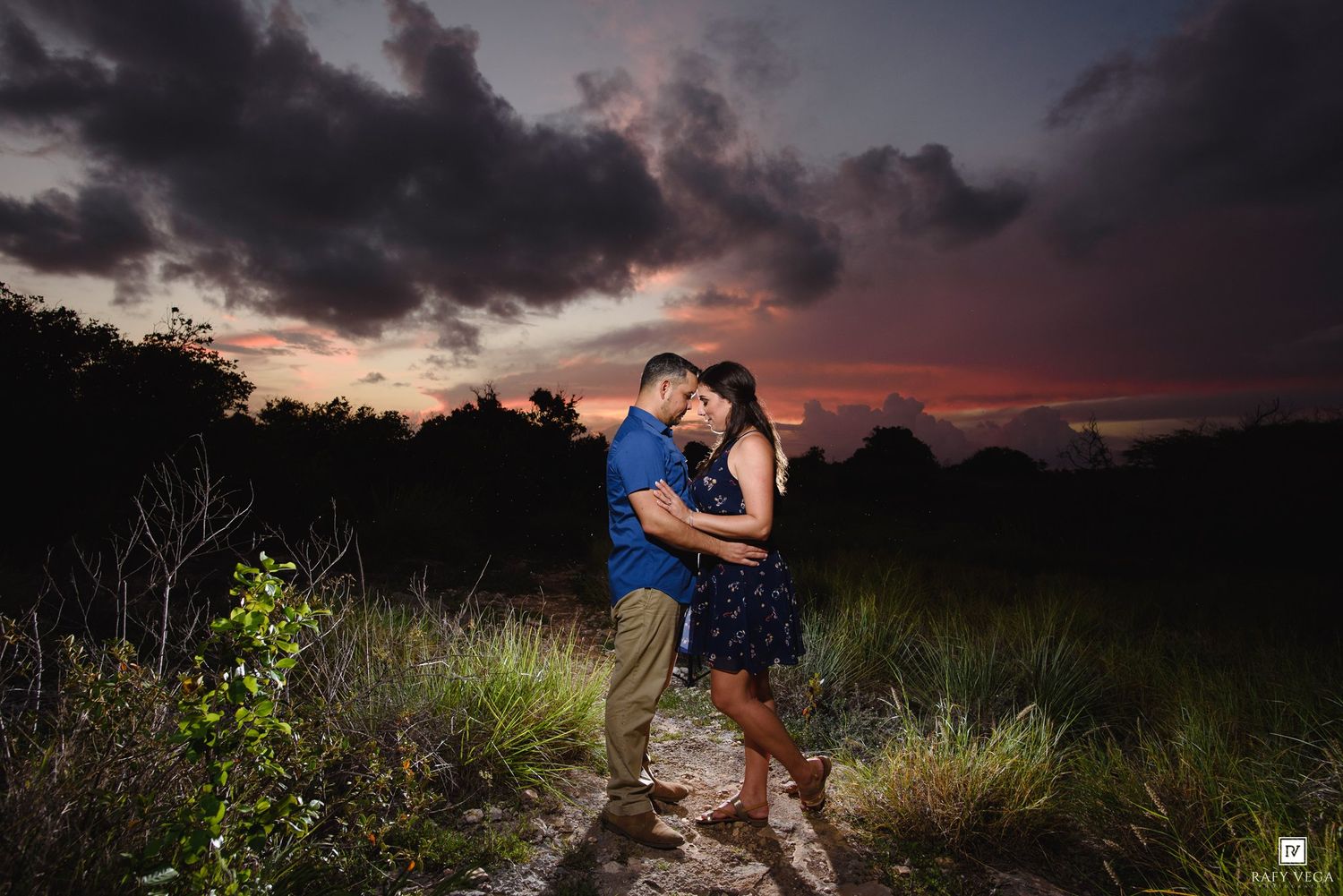 Couple embracing romantically at sunset with dramatic colorful sky and lush greenery in background.