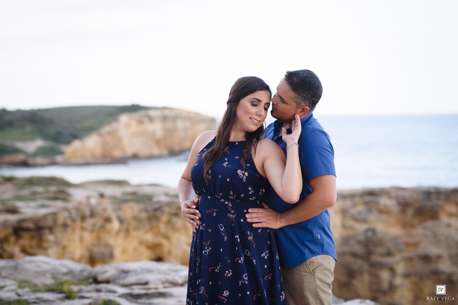 Couple sharing a tender moment on rocky seaside cliffs, man whispering to woman in navy floral dress.