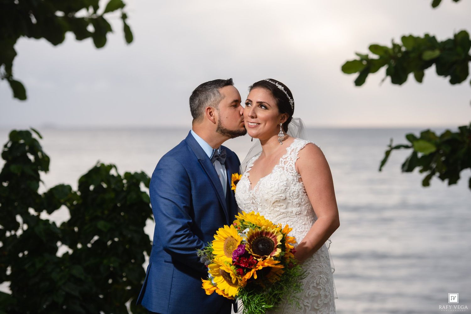 Romantic bride and groom sharing a kiss by the ocean at sunset, bride holding a vibrant sunflower bouquet.