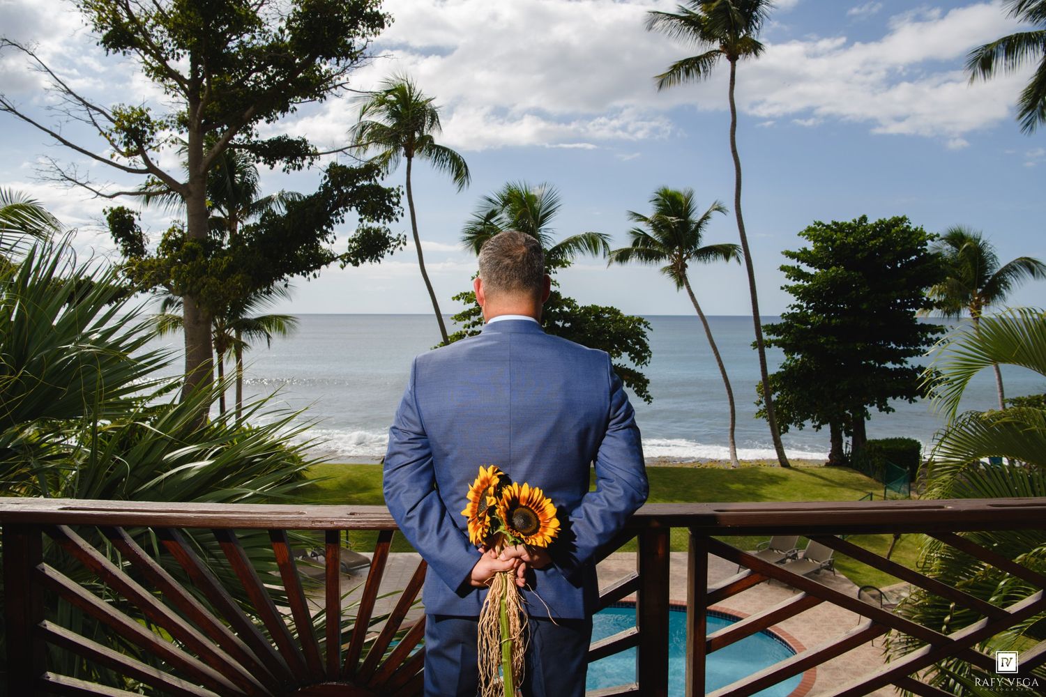 Groom in blue suit holds sunflowers on tropical balcony overlooking palm trees and ocean at destination wedding.