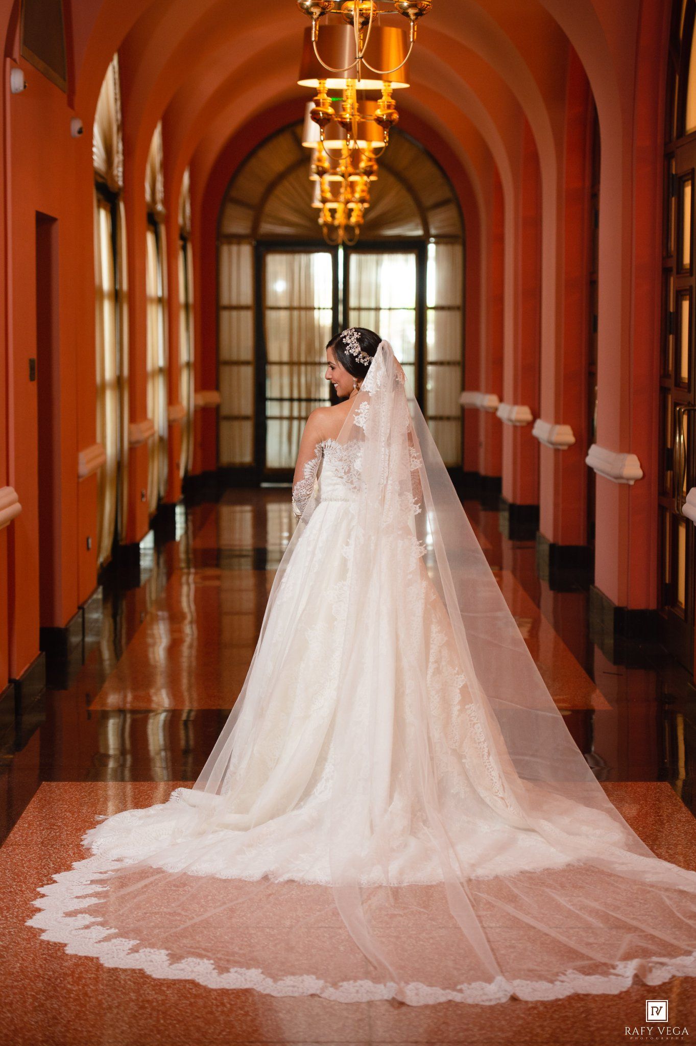 Bride in stunning white ball gown with long cathedral veil standing in ornate arched hallway.