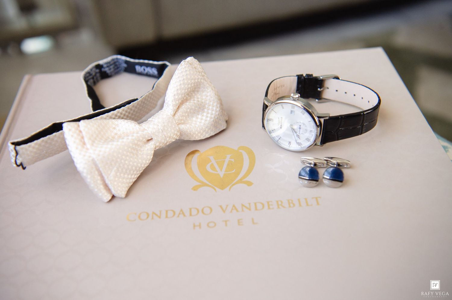 White bow tie, classic watch, and blue cufflinks displayed on a Condado Vanderbilt Hotel box for a wedding.