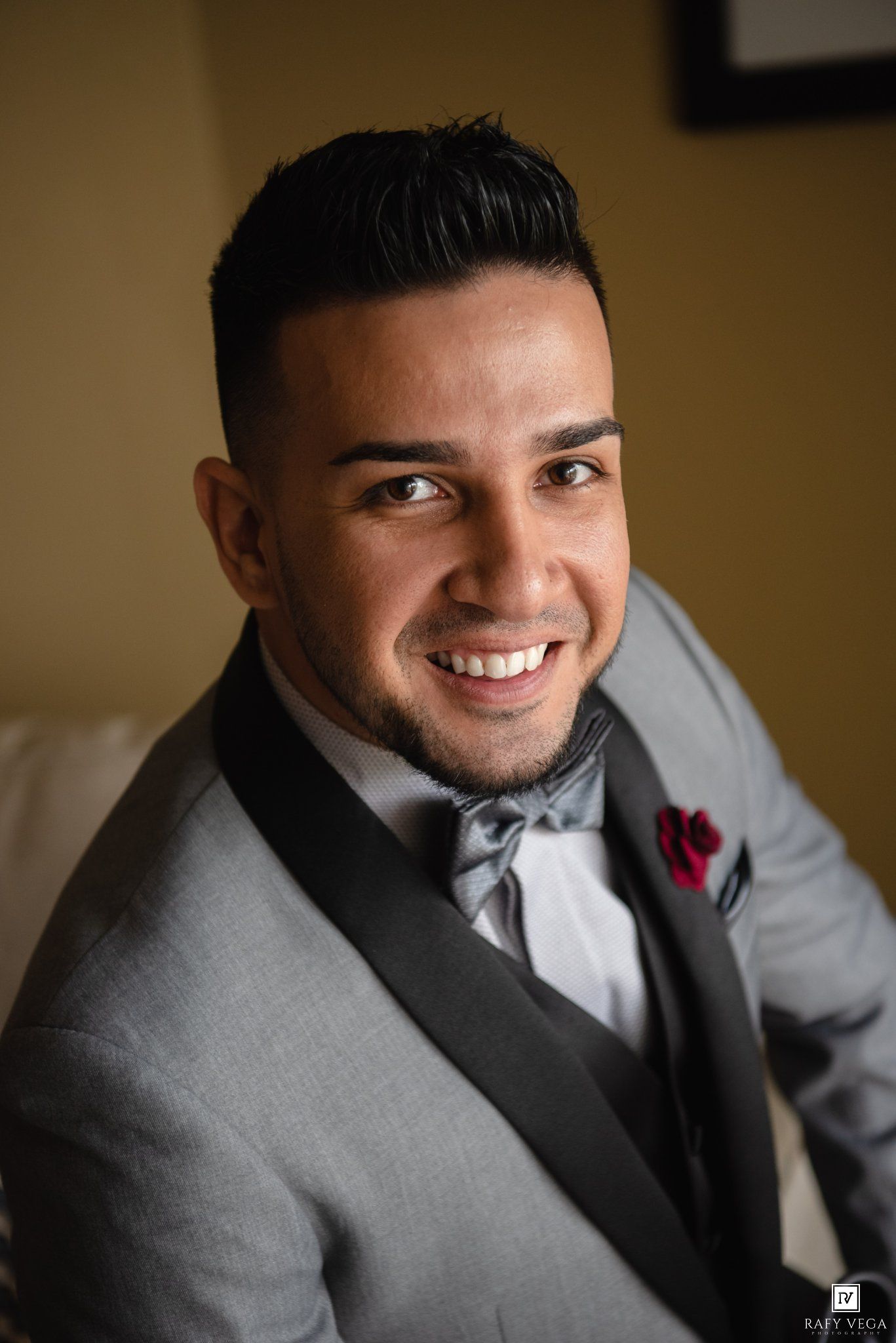 Smiling groom in gray tuxedo with bow tie and red pocket square posing for wedding portrait.