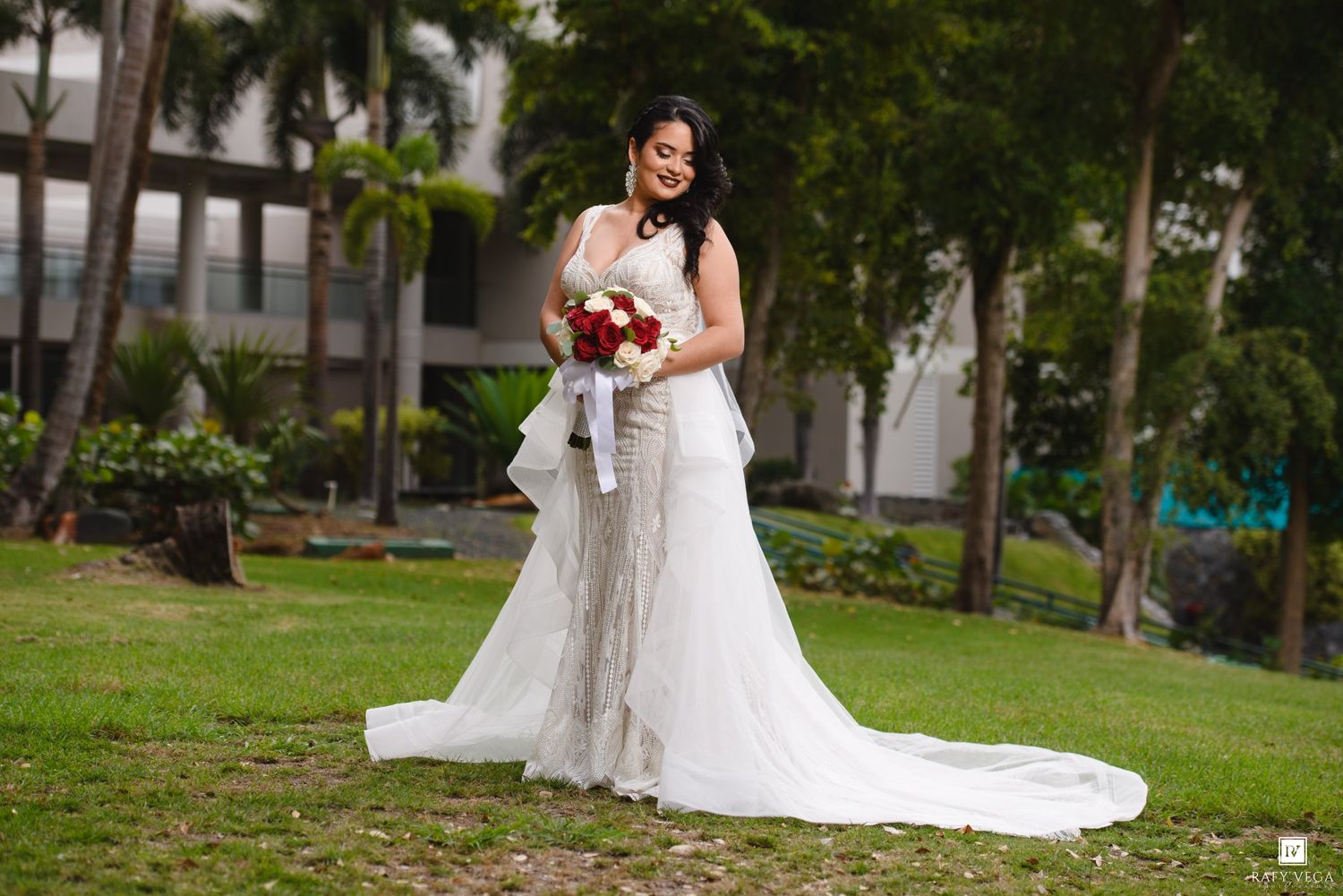 Bride in white lace wedding gown with train holds red bouquet outdoors on green lawn.