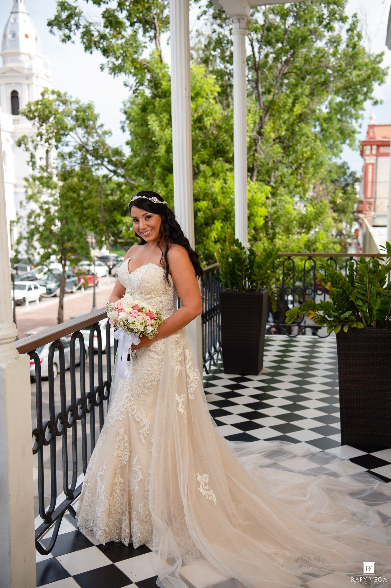 Bride in lace wedding gown holding bouquet on checkered balcony with white columns and green trees.