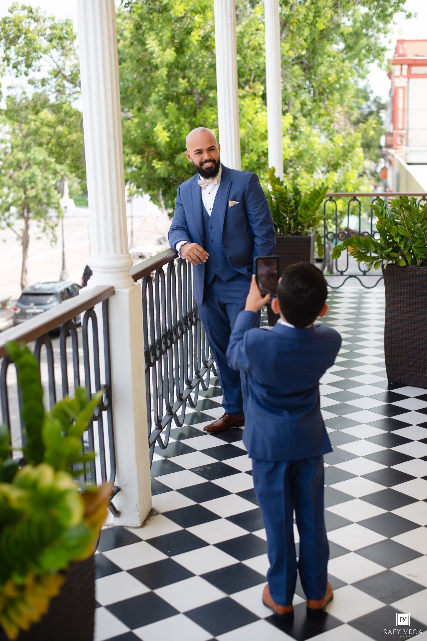 Man in blue suit smiling at a boy in matching suit on a checkered balcony.