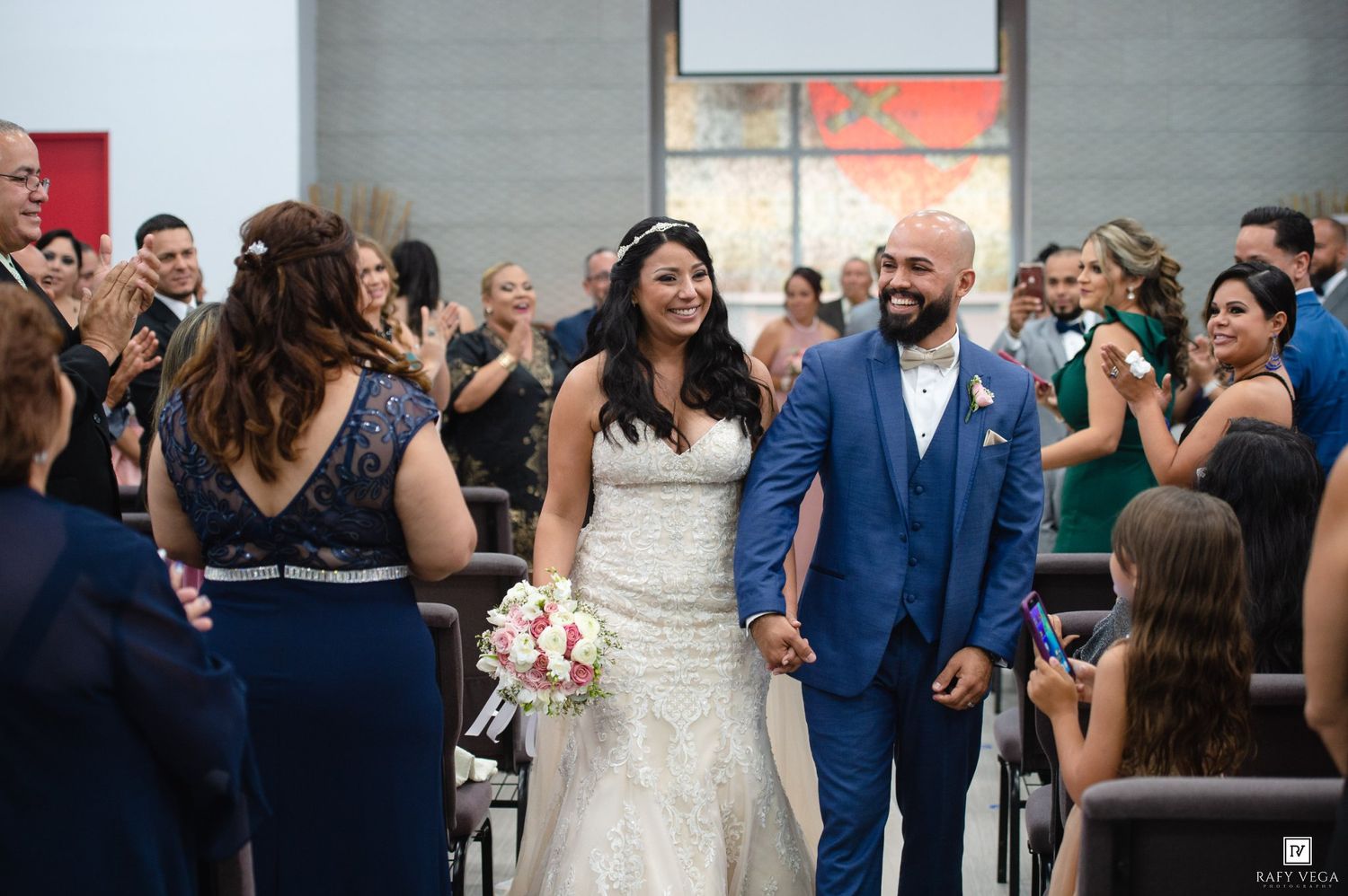 Smiling newlywed couple walks down the aisle hand-in-hand at their wedding ceremony surrounded by applauding guests.