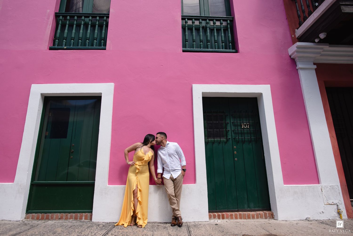 Couple sharing a romantic moment against a vibrant pink building with green doors in Old San Juan, Puerto Rico.