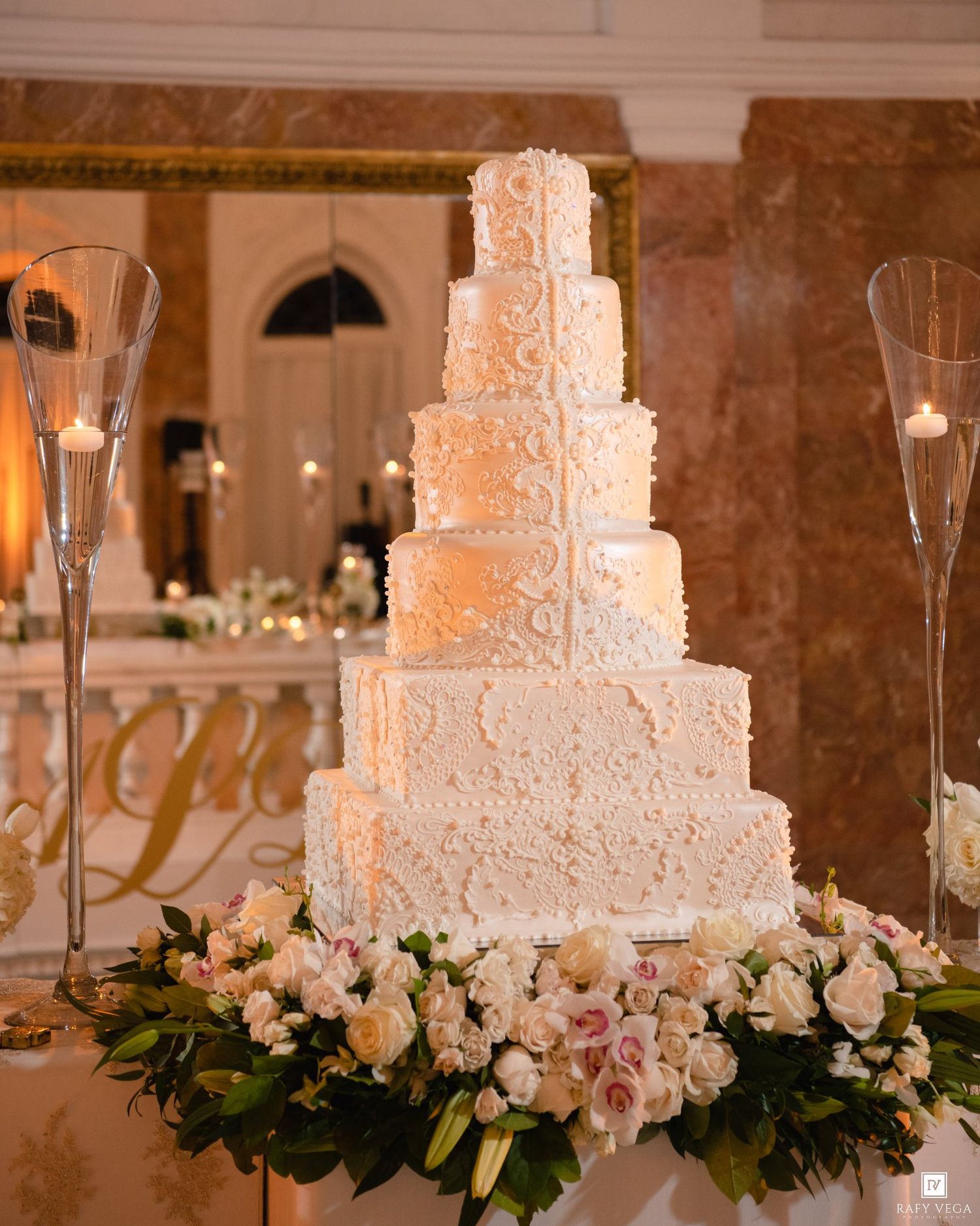 Elegant six-tier white wedding cake adorned with lace details, surrounded by pink and white flowers on a decorated table.