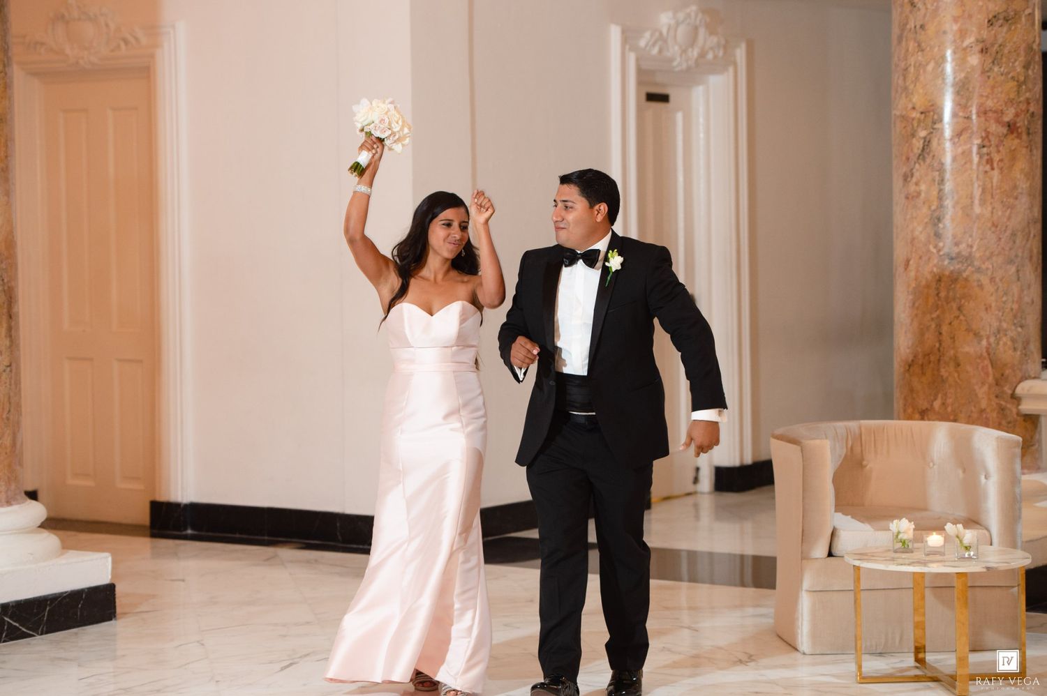 Joyful bride in sleek gown raises bouquet as groom in tuxedo escorts her into elegant reception hall.