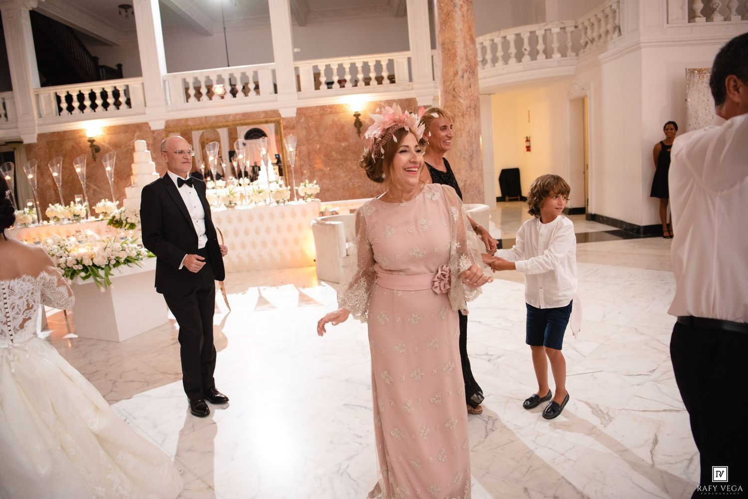 Elegant wedding reception in a grand ballroom with guests dancing, featuring floral arrangements and marble floors.