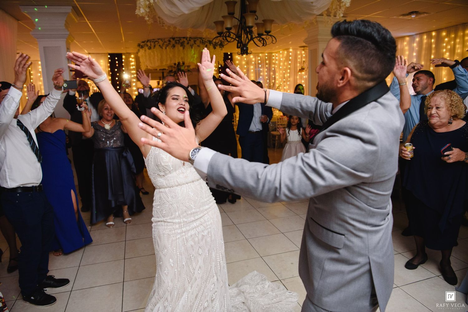 Bride and groom dancing joyfully with hands raised at elegant wedding reception with guests celebrating.