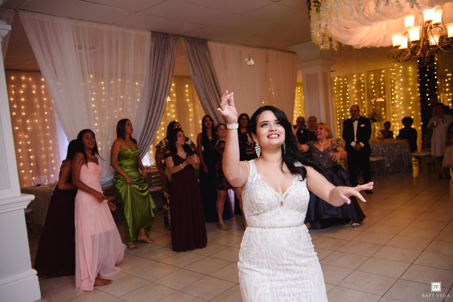 Bride in white lace gown tossing bouquet at elegant wedding reception with guests and fairy lights.