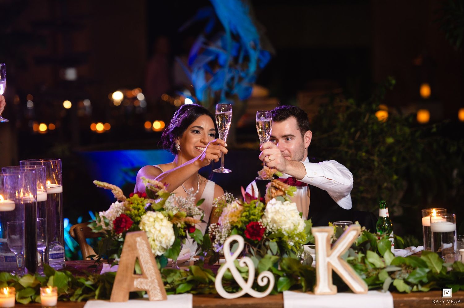 Bride and groom toasting with champagne flutes at wedding reception sweetheart table decorated with floral centerpiece and candles.