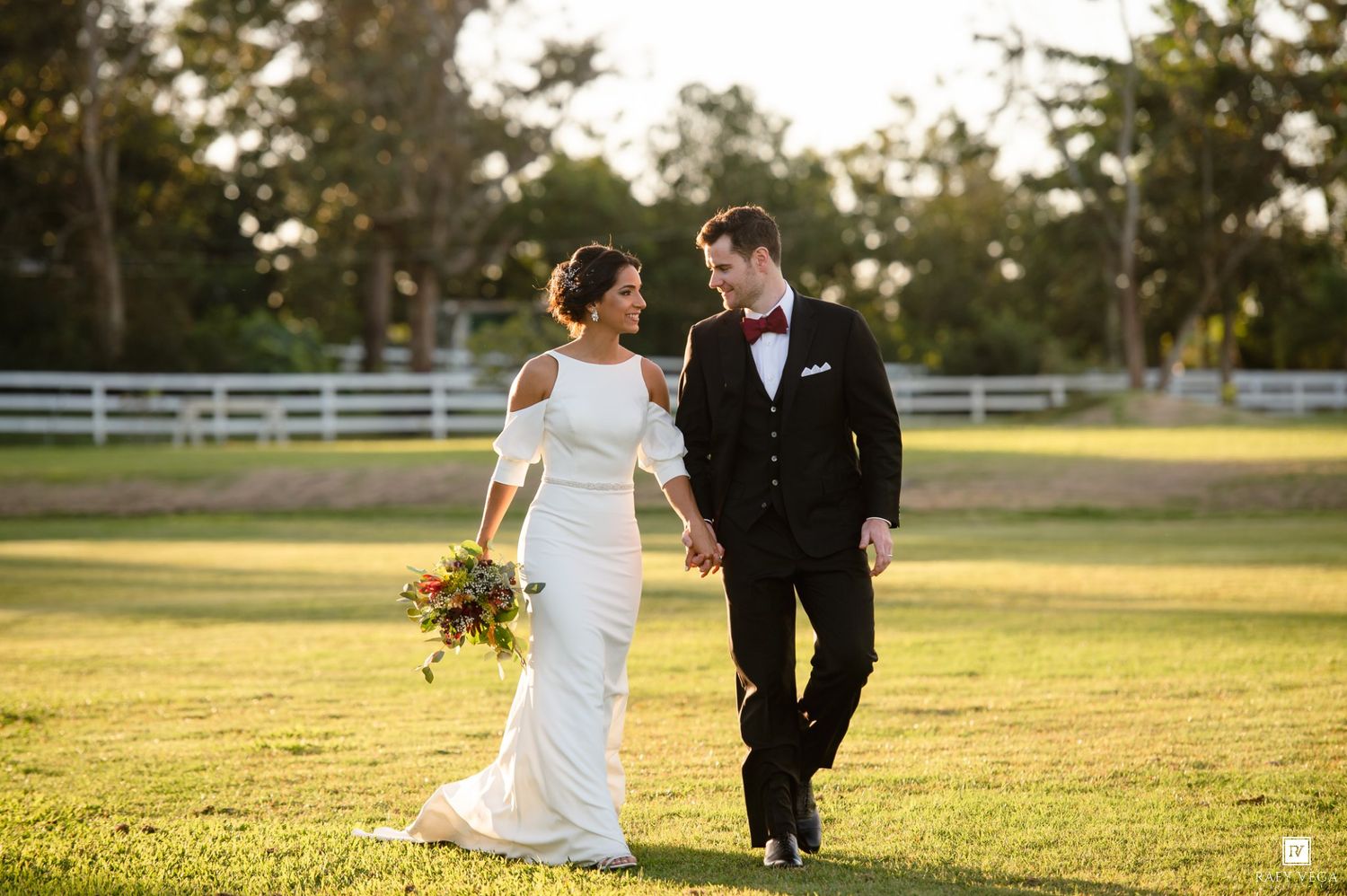 Bride and groom walking hand in hand on lush green lawn at golden hour outdoor wedding venue.