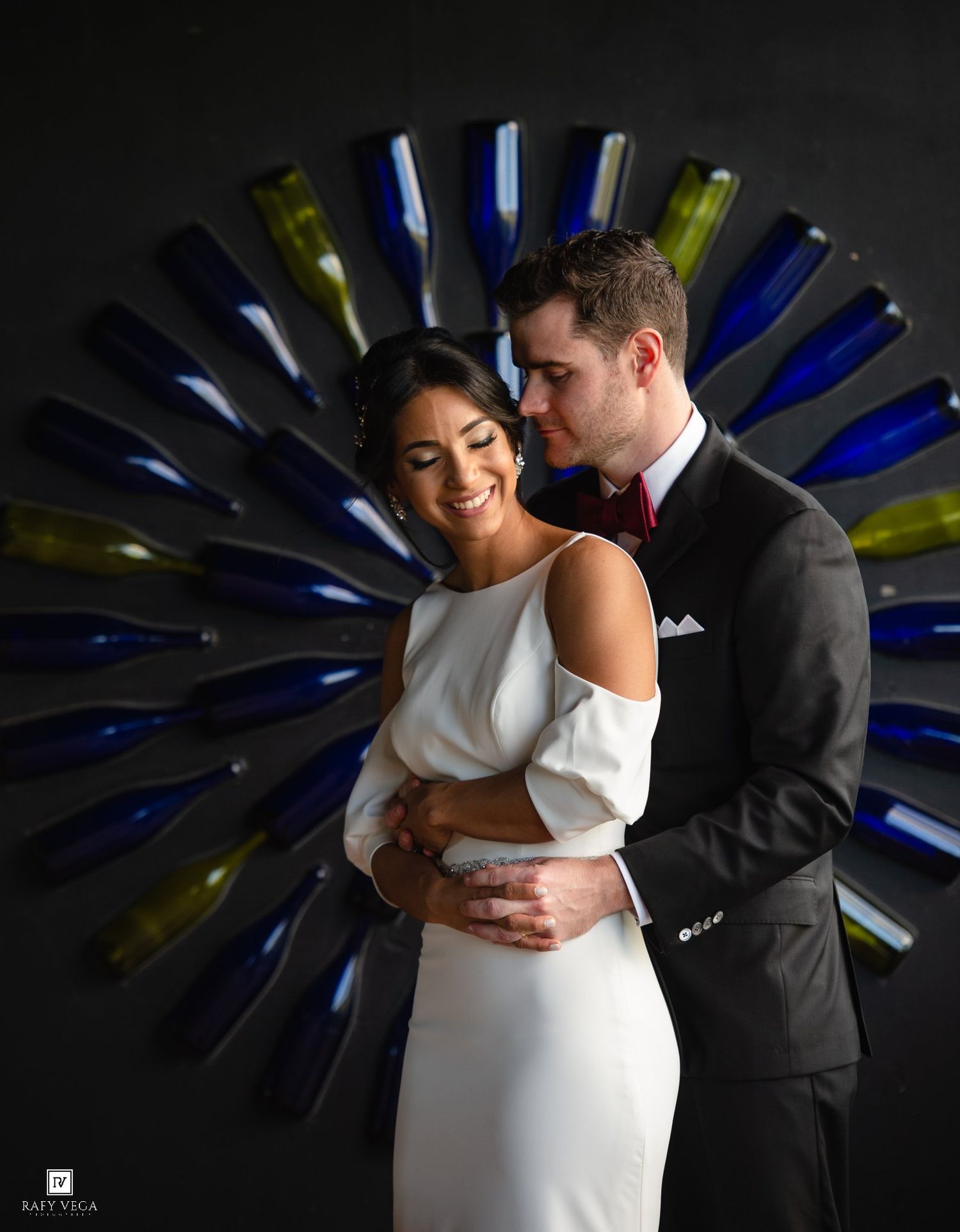 Bride and groom embrace before a decorative wine bottle wall art display in a romantic wedding portrait.